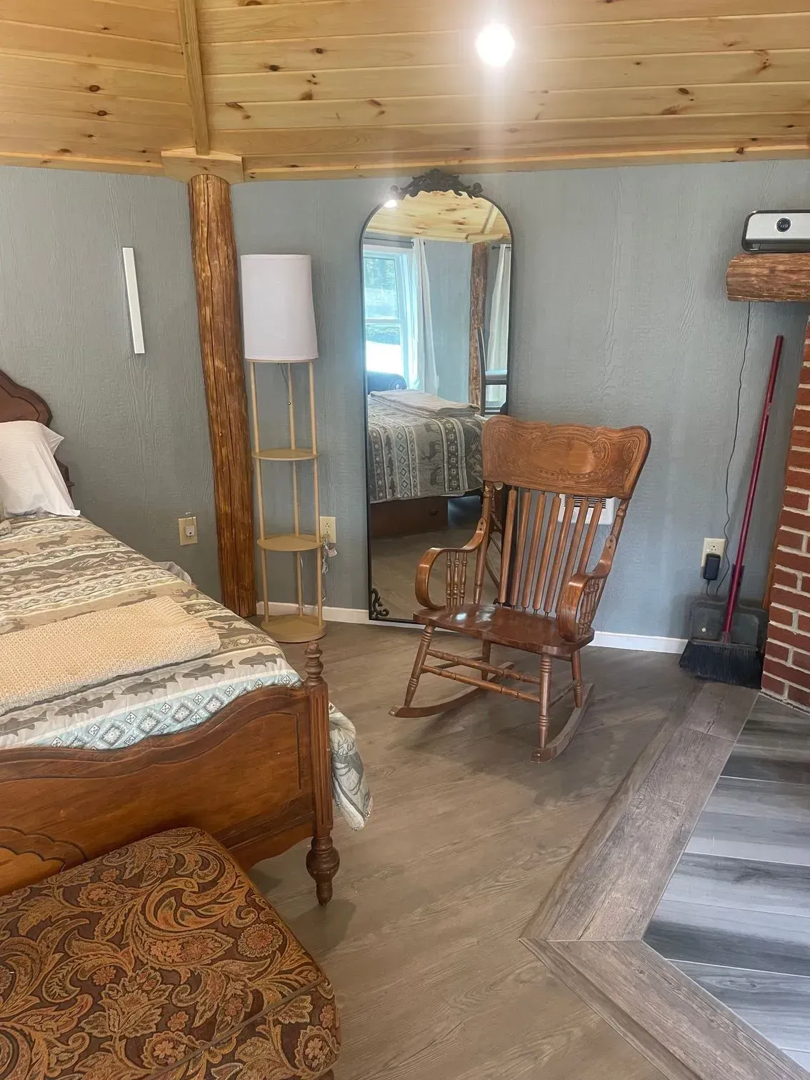 A rustic bedroom with a wooden bed, a rocking chair, an arched floor mirror, and a brick fireplace.