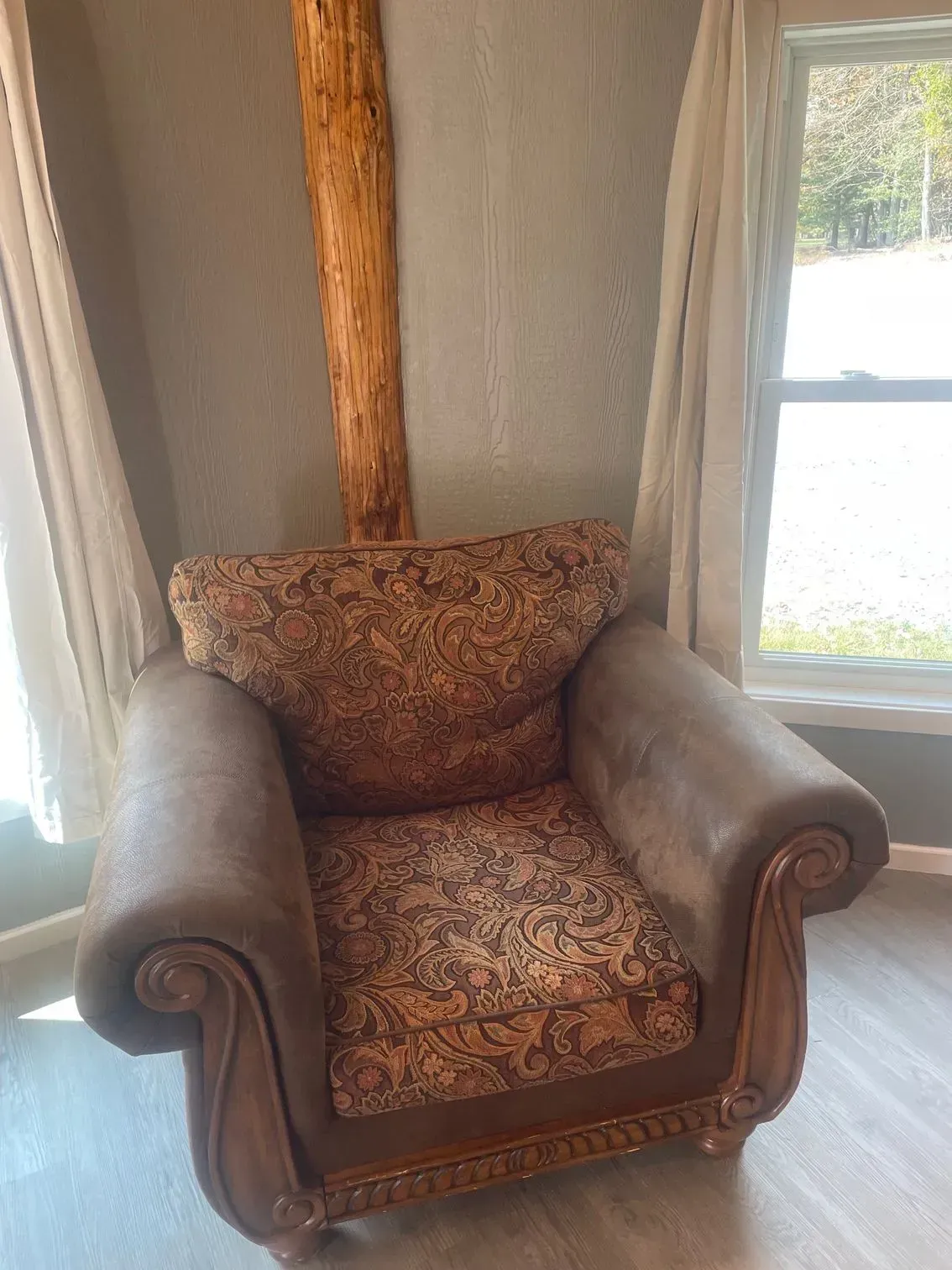 A brown, upholstered armchair with a patterned paisley cushion, positioned in a corner near a window and wooden post.
