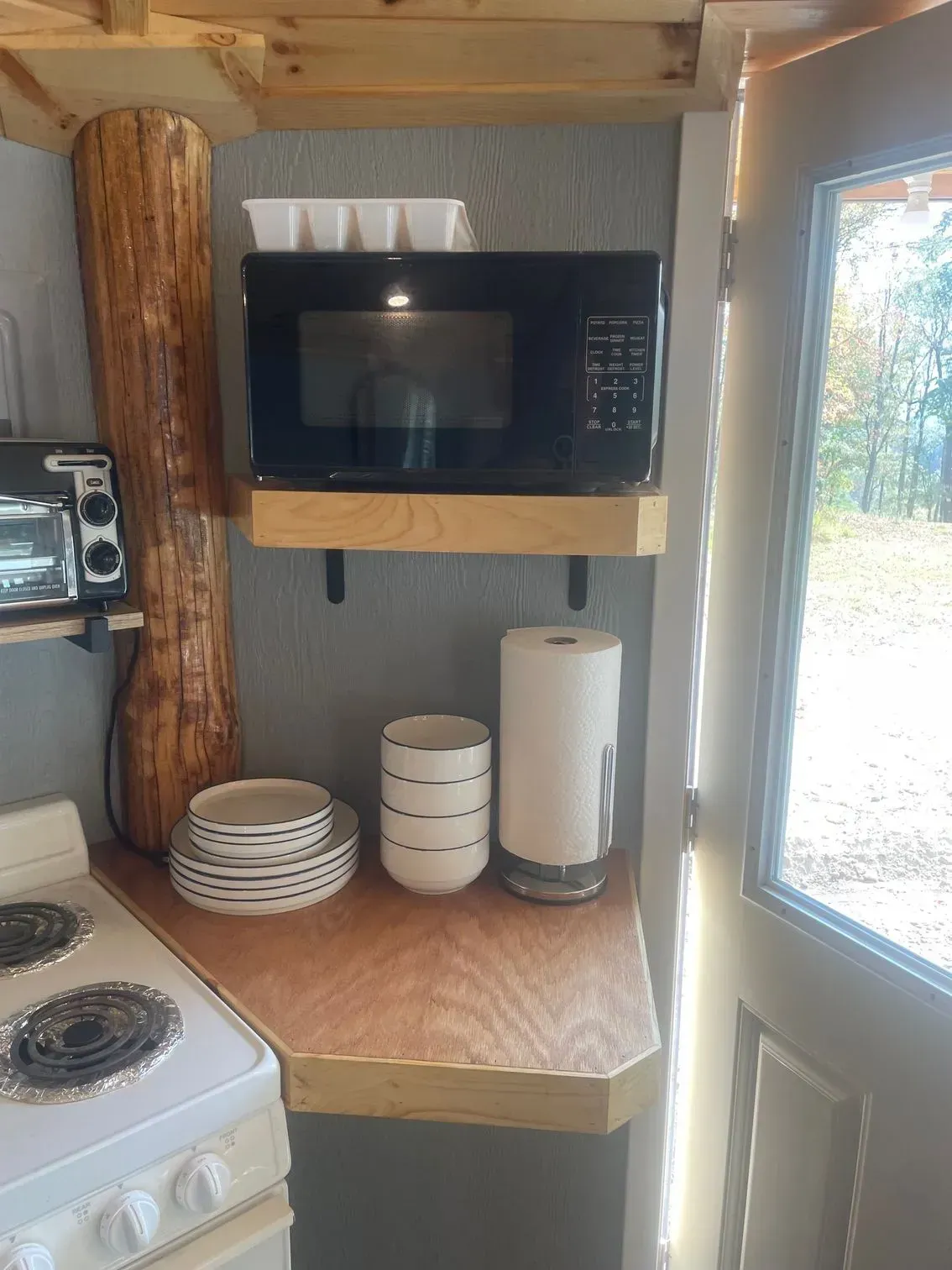 A wooden kitchen counter with a microwave on a shelf above, next to a stove, stacks of bowls, and a paper towel roll.