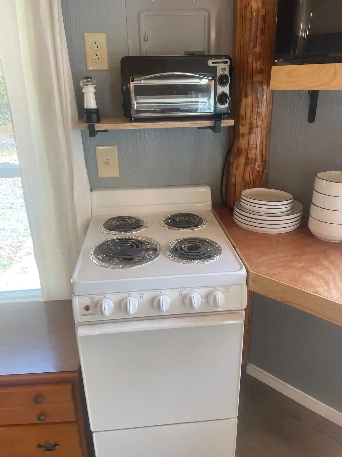 A white electric stove sits in a kitchen corner with a toaster oven on a shelf above it and a stack of plates nearby.