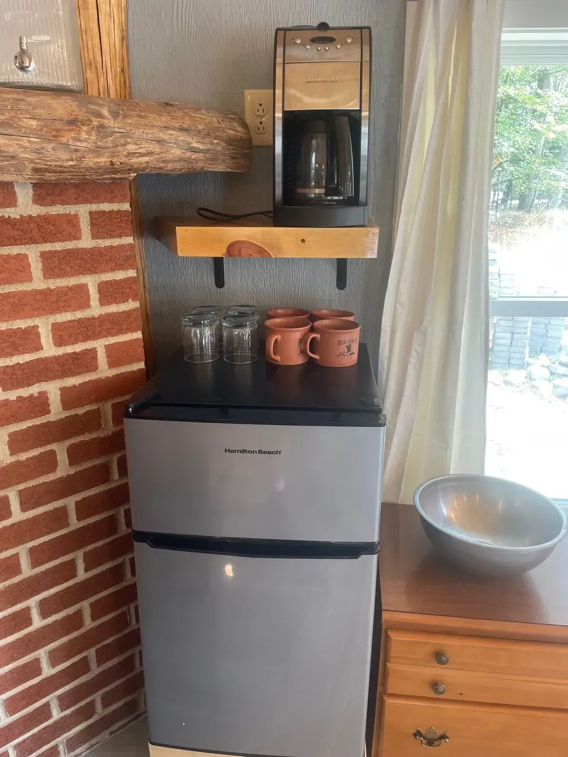 A mini-fridge with mugs and glasses on top sits below a shelf holding a coffee maker against a brick wall.