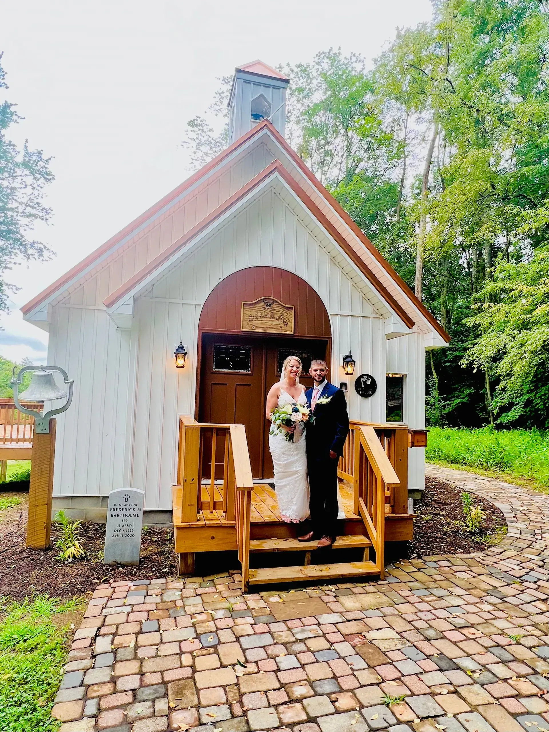 A bride and groom standing on the wooden steps of a small, white chapel set against a background of green trees.