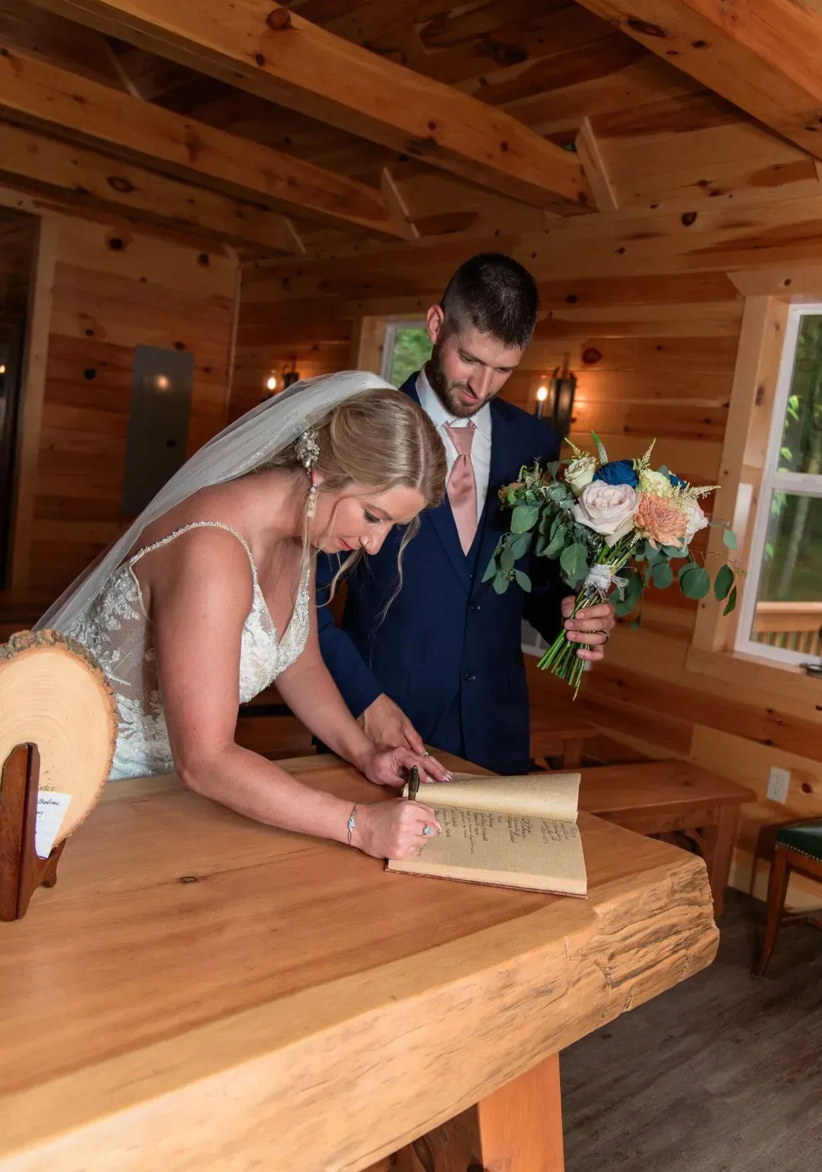 A bride in a white wedding gown and a groom in a navy suit sign a guestbook at a rustic wooden table inside a cabin.