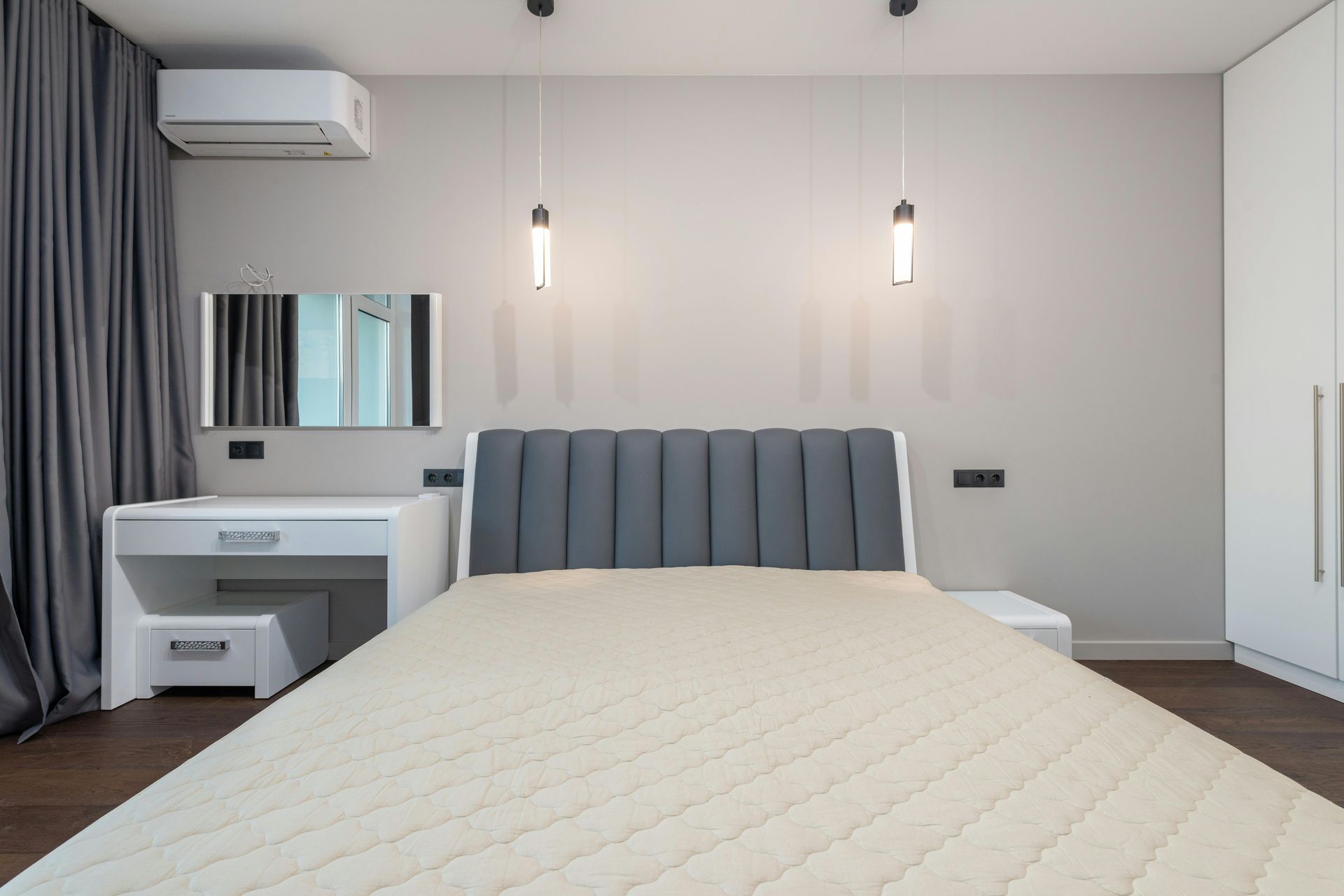 Modern bedroom with a white desk, grey upholstered headboard, beige mattress, and grey curtains against a light wall.