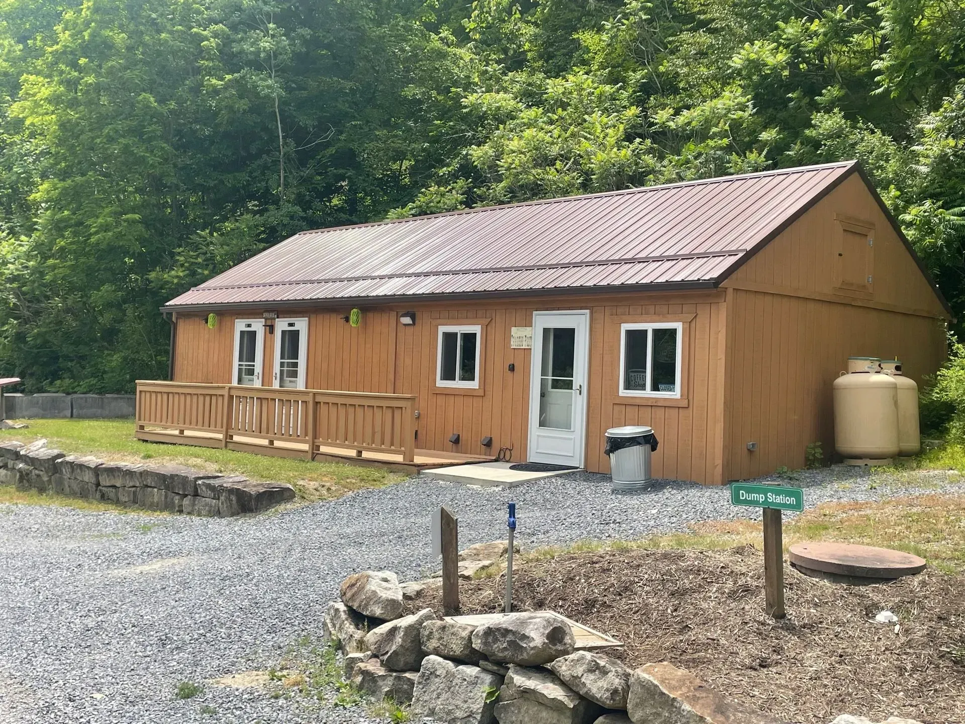 Small tan cabin with porch in a wooded campsite, beside gravel and a rock fire ring.