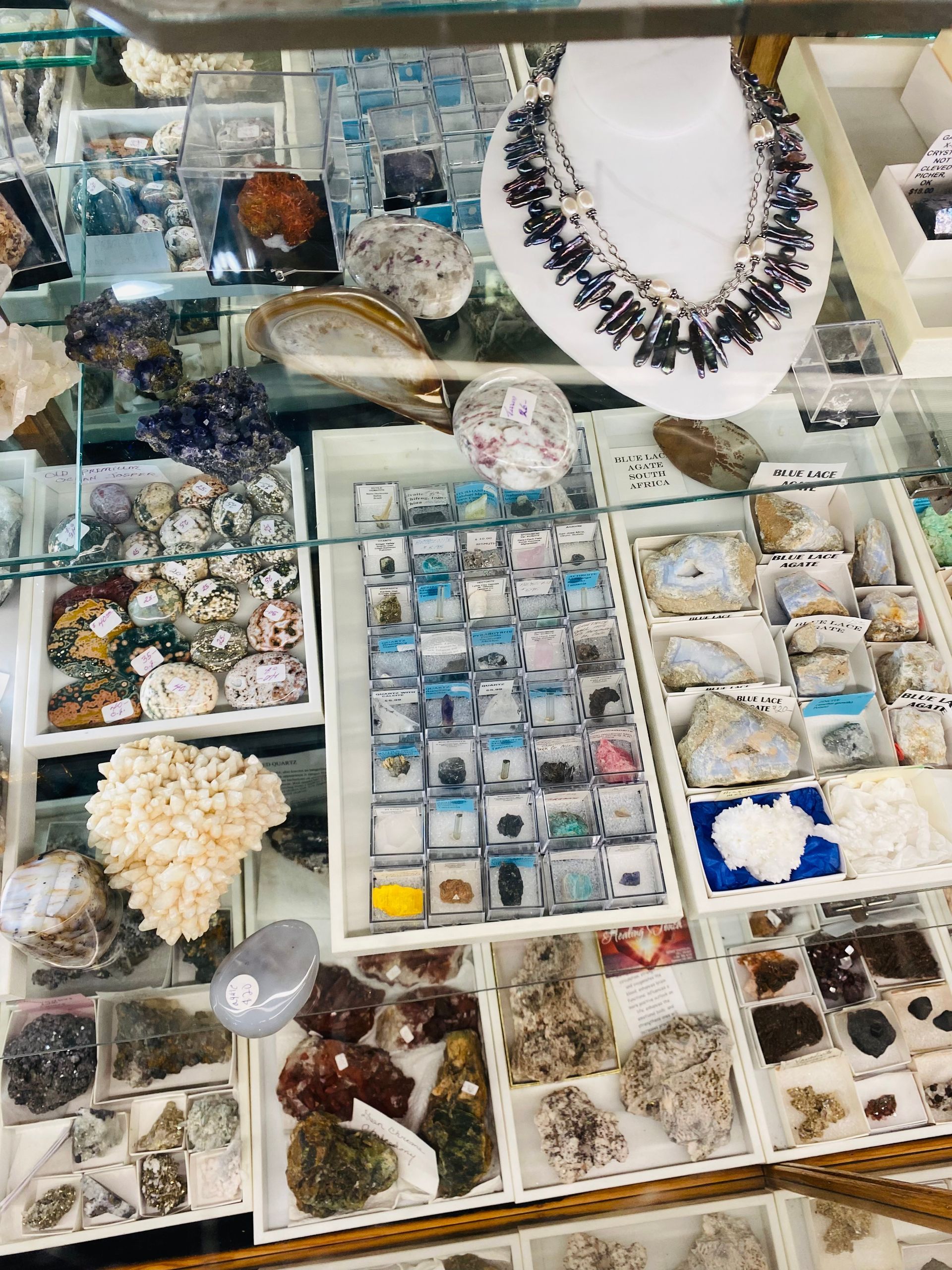 Display of assorted crystals and gemstone jewelry in white trays, with a black stone necklace on a bust.