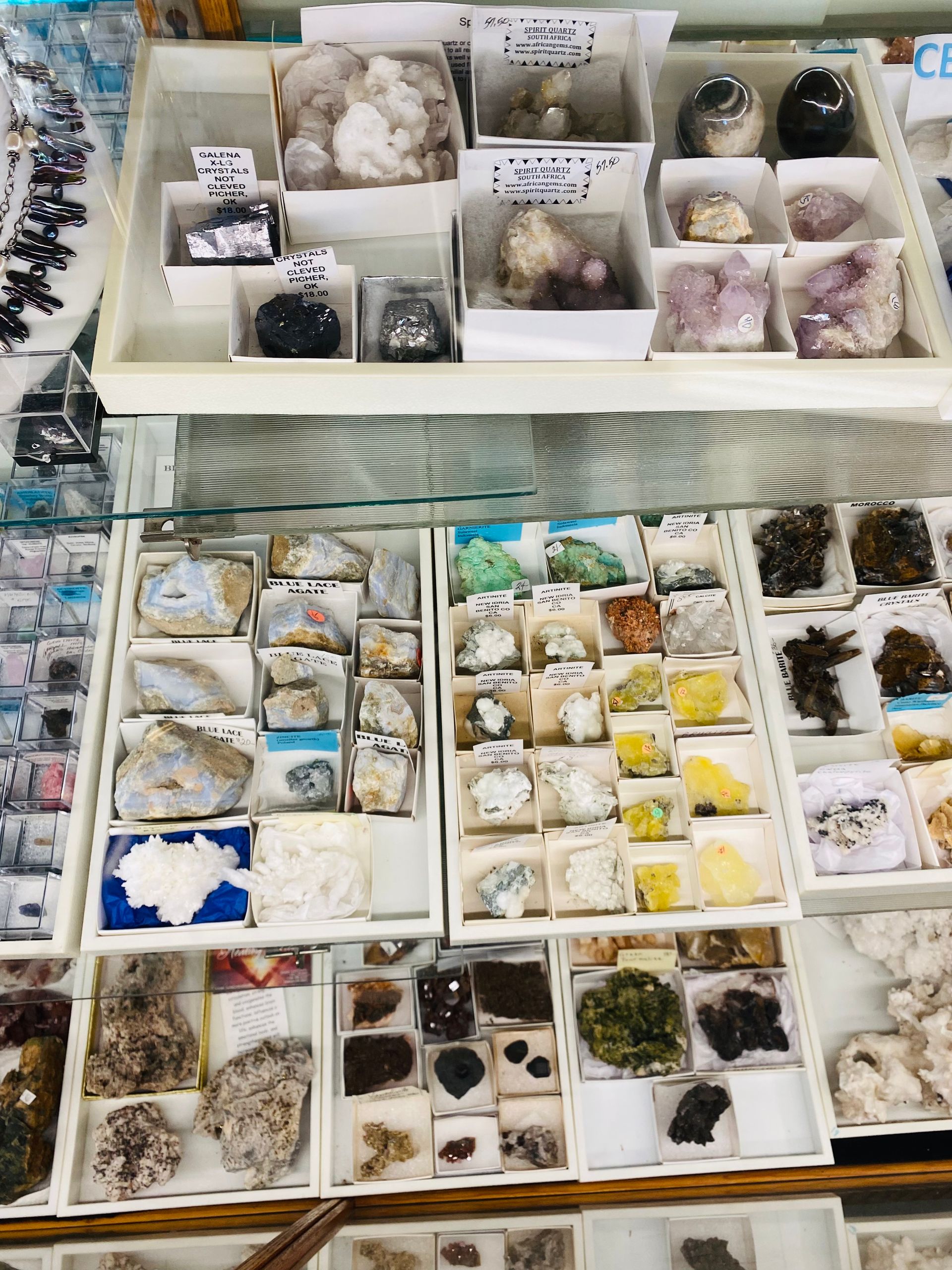 Tray display of assorted crystal and mineral specimens in small white compartments, viewed from above.