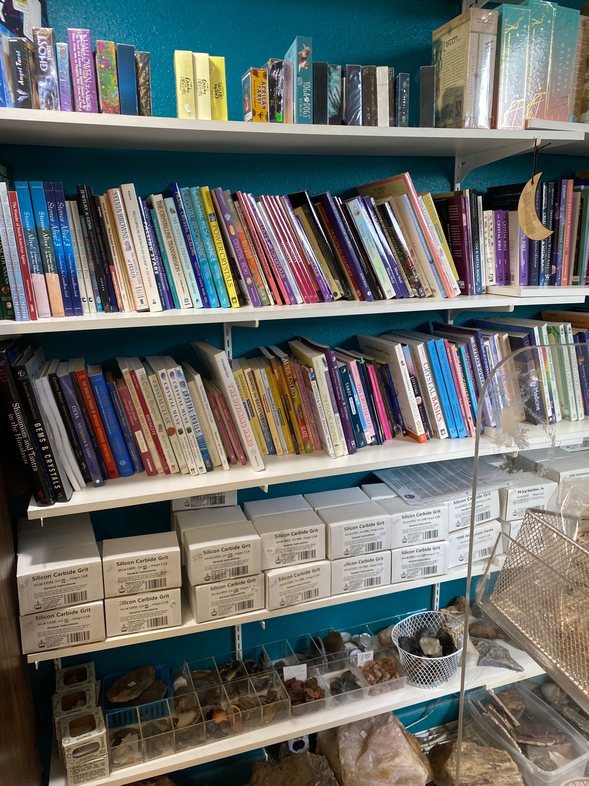 Colorful books stacked on angled white shelves in a shop, with boxes and craft supplies below.