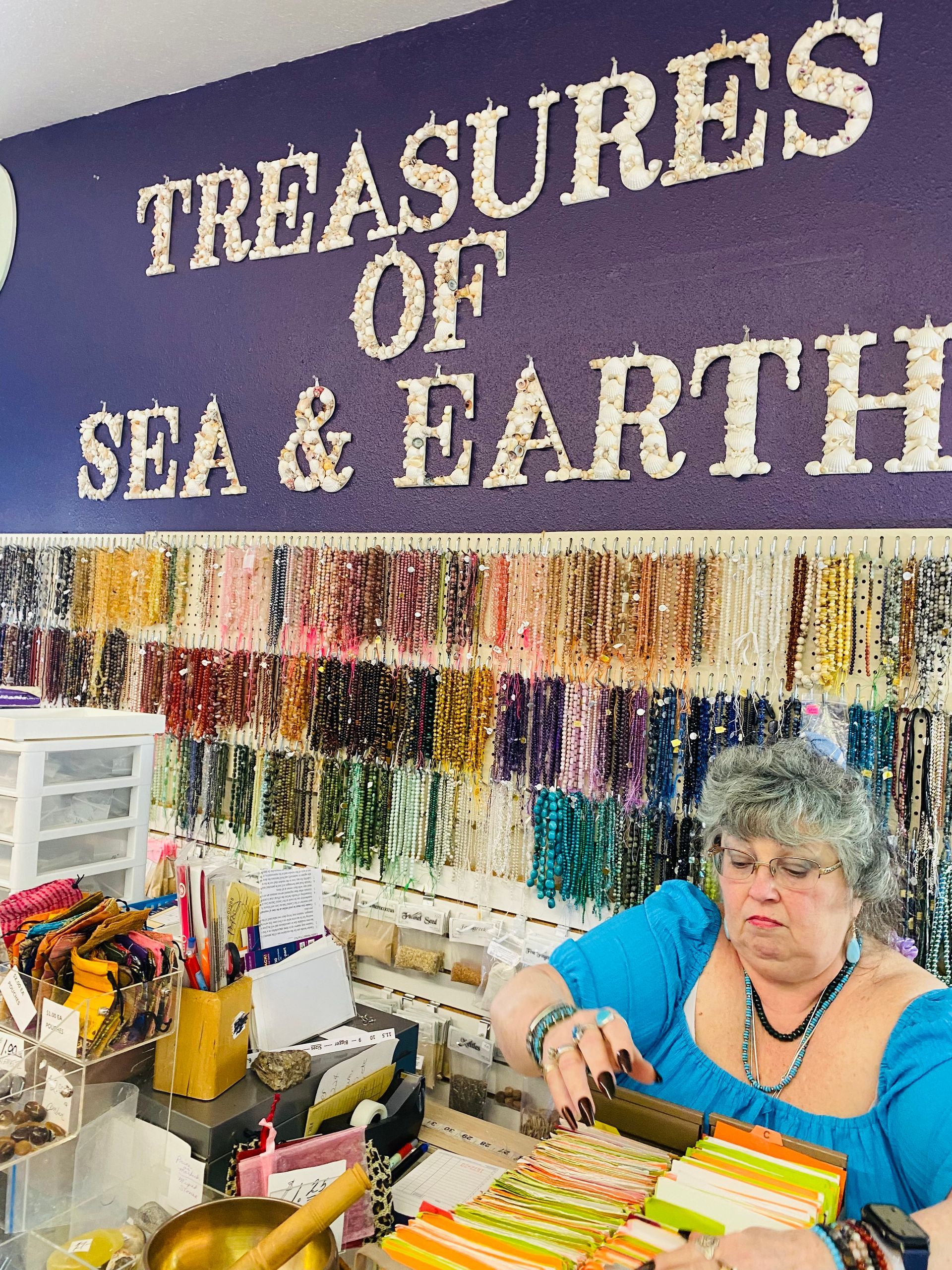 Vendor booth with “Treasures of Sea & Earth” sign, colorful bead strands, and a woman seated at a craft table