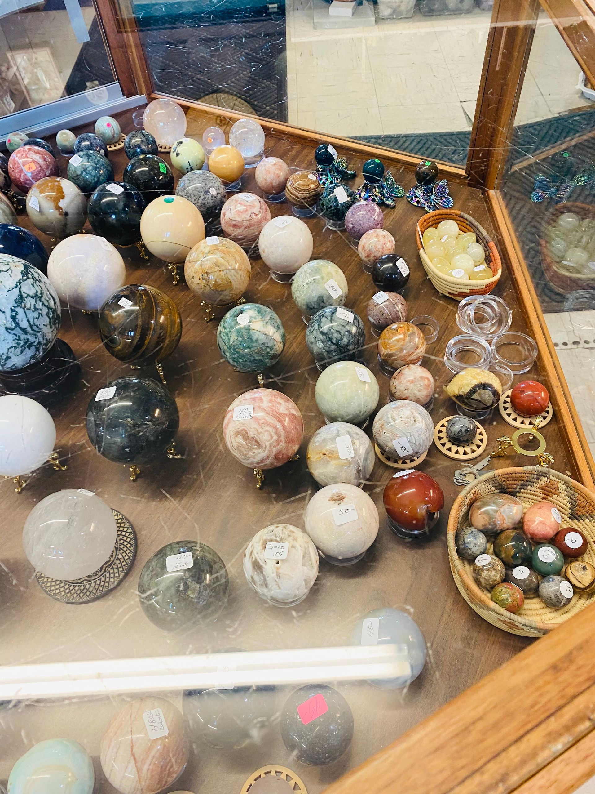 Assorted polished gemstone spheres displayed in glass cases, with small stone eggs and carvings on a wooden tray.