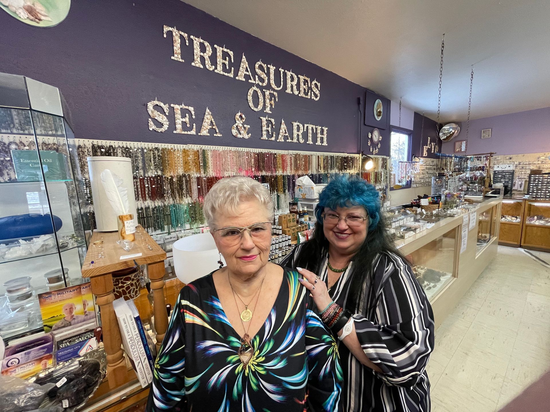 Two women smiling in a sea-themed shop with shelves of souvenirs and a “Treasures of Sea & Earth” sign.