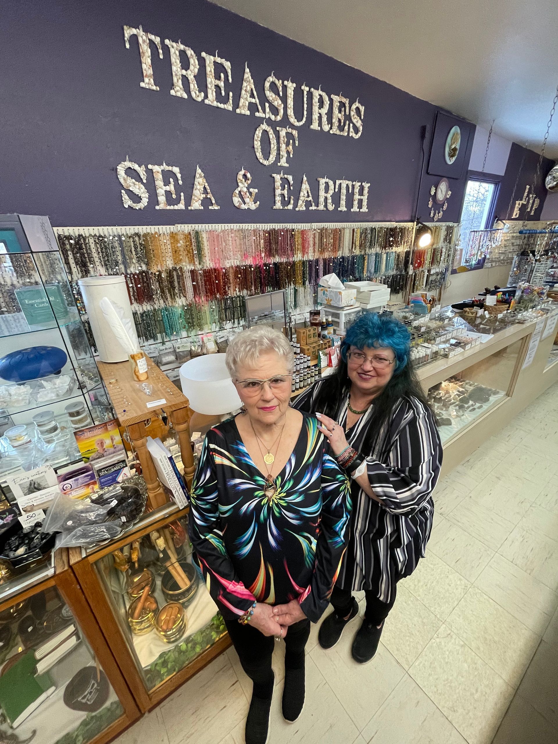 Two women posing inside a shop called “Treasures of Sea & Earth,” with jewelry and gifts on display.
