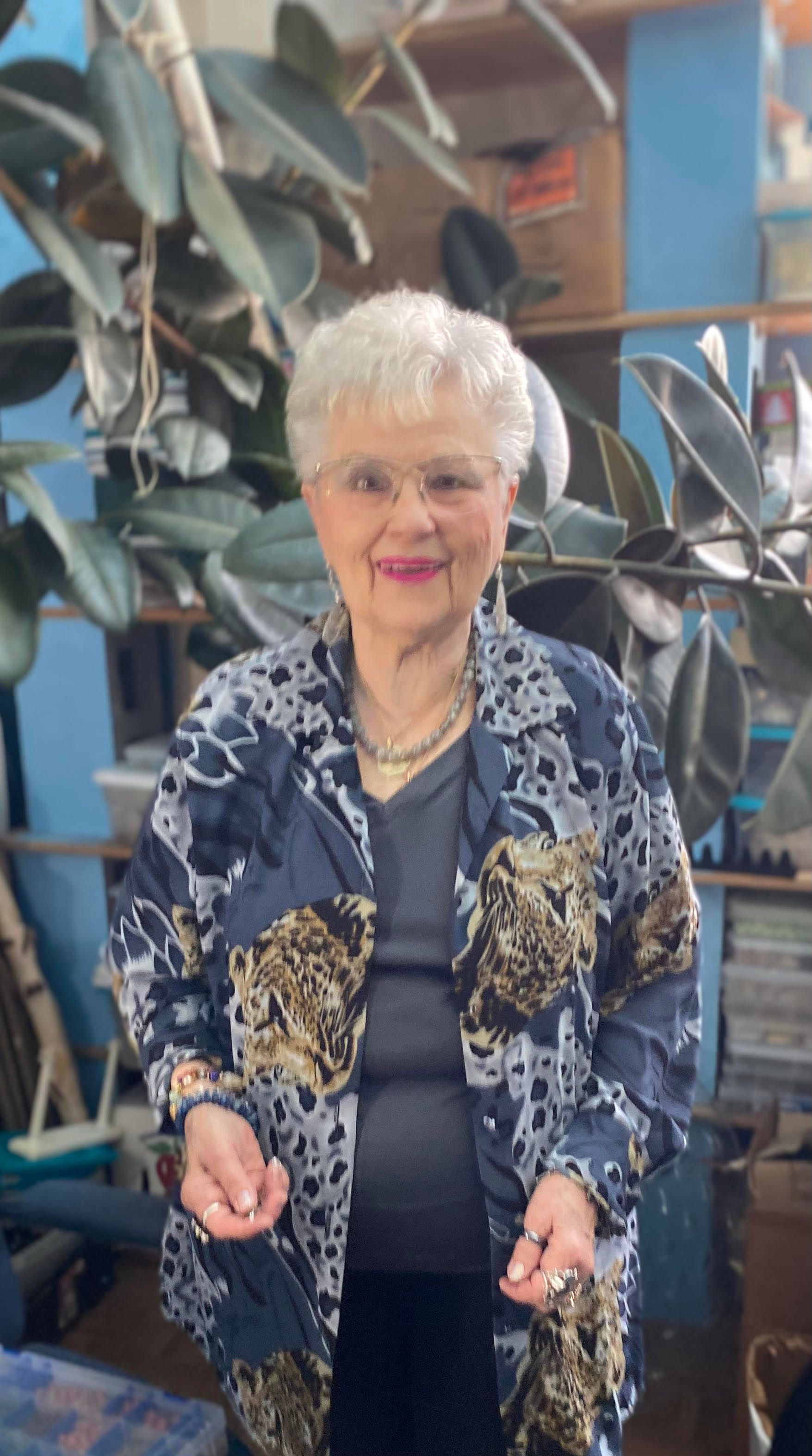 Woman standing indoors among large plant leaves, wearing a blue patterned jacket and smiling.