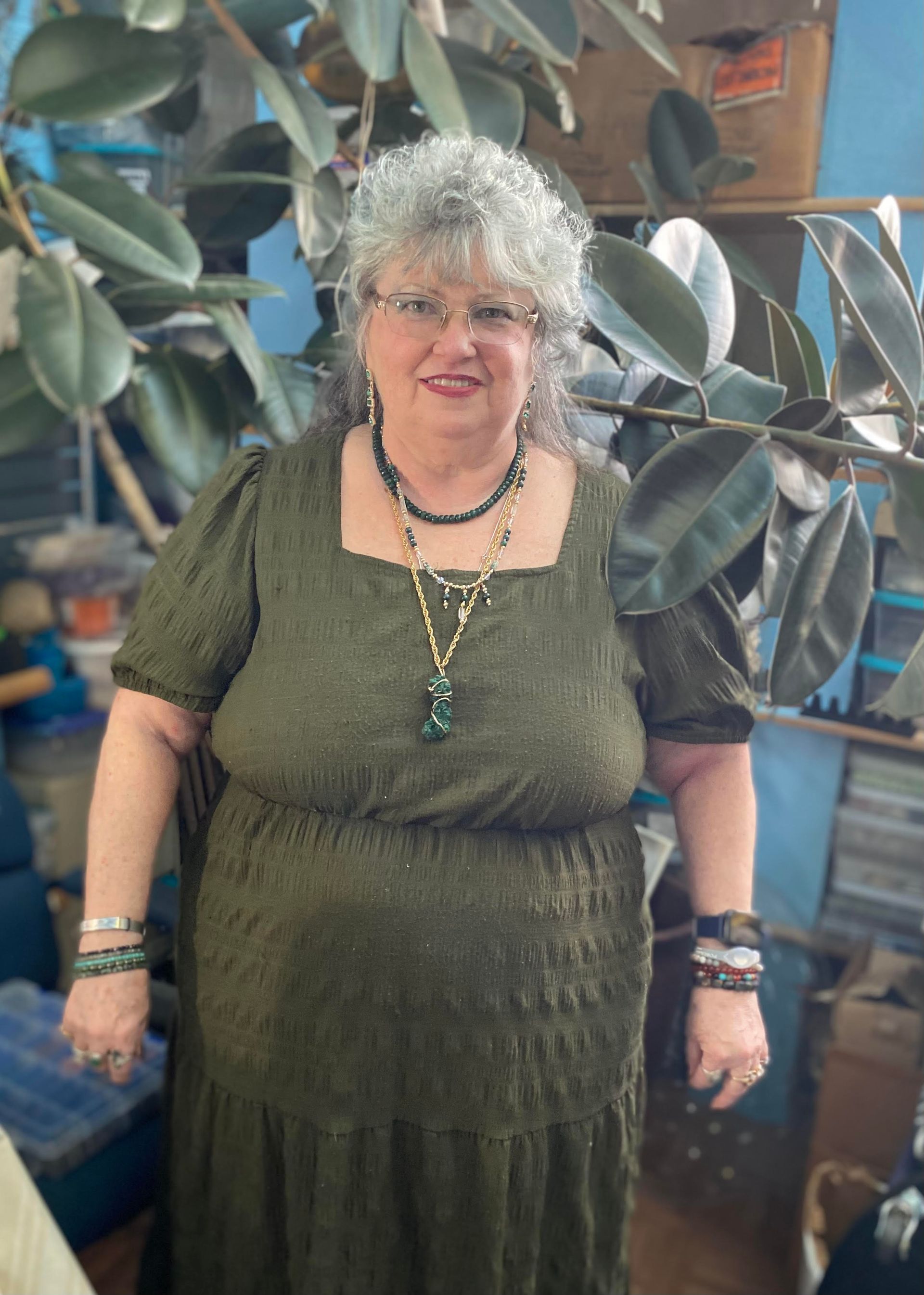 Woman in an olive-green dress and jewelry standing indoors among large plant leaves.
