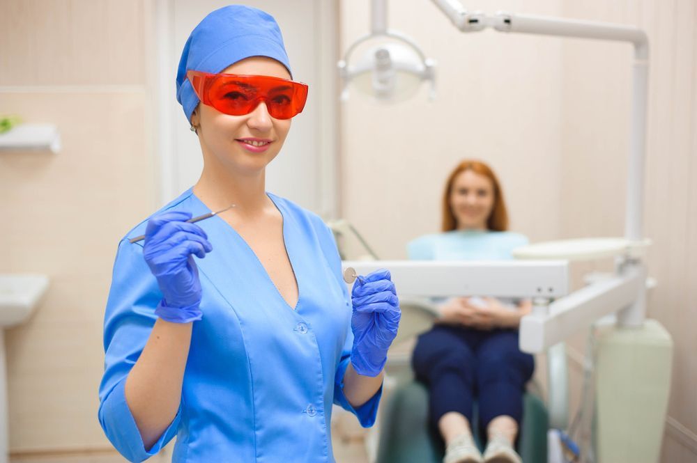 Dentist in blue scrubs and protective glasses, holding tools, smiling, with patient in background.