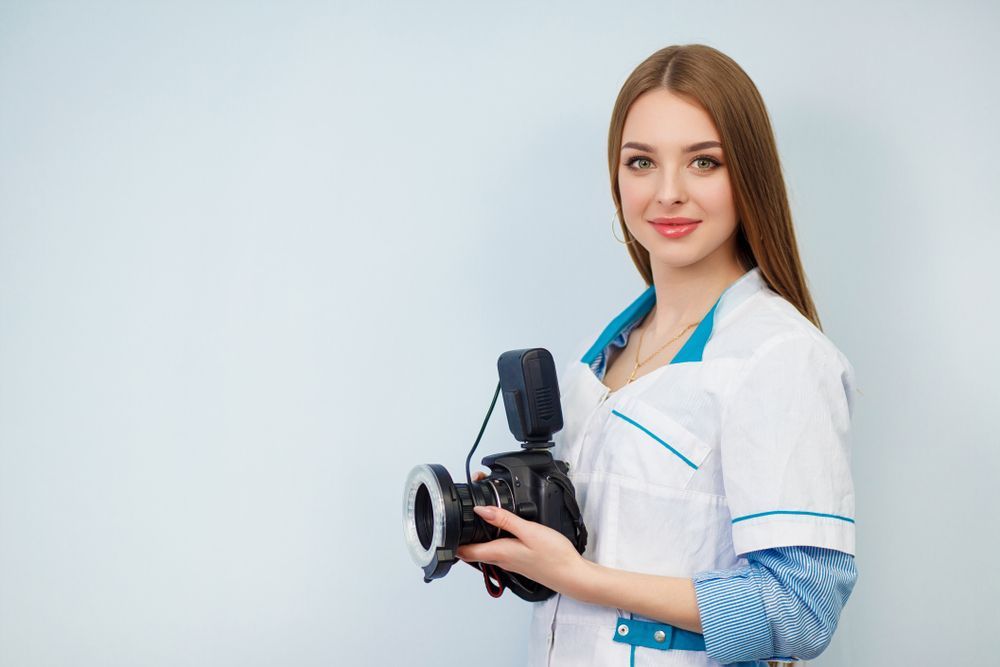 Woman in white lab coat holding a camera with flash, smiling. Light blue background.