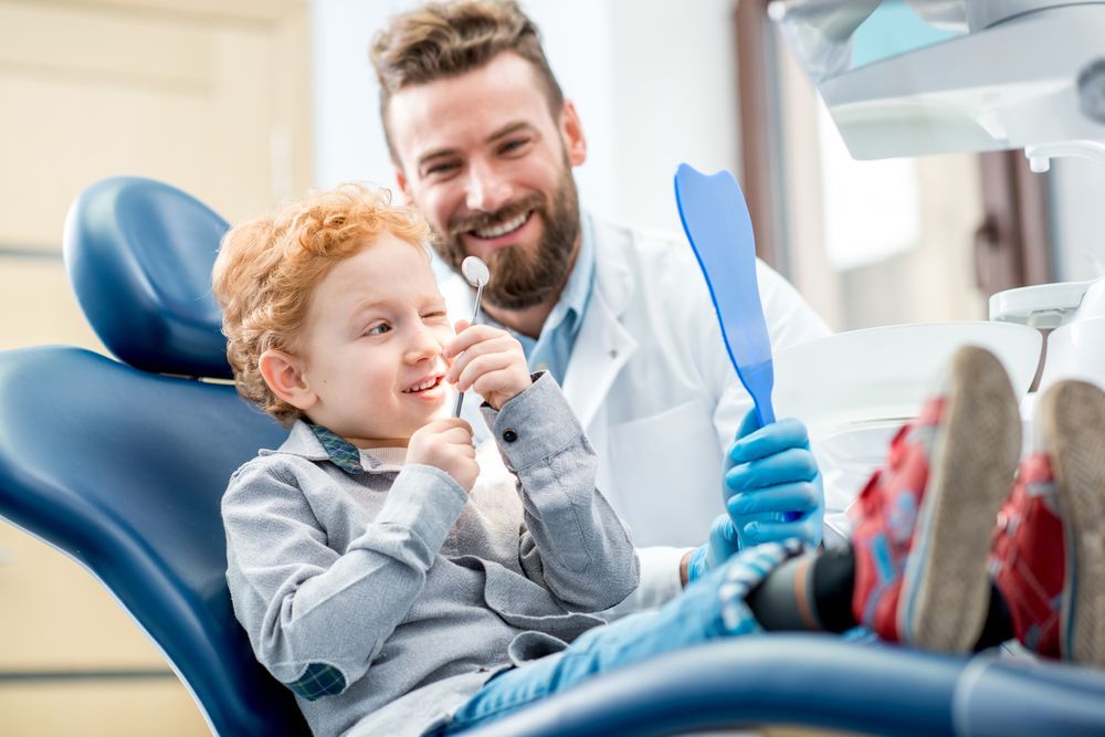 A child in a dentist chair looking at their teeth in a mirror, smiling at a smiling dentist.
