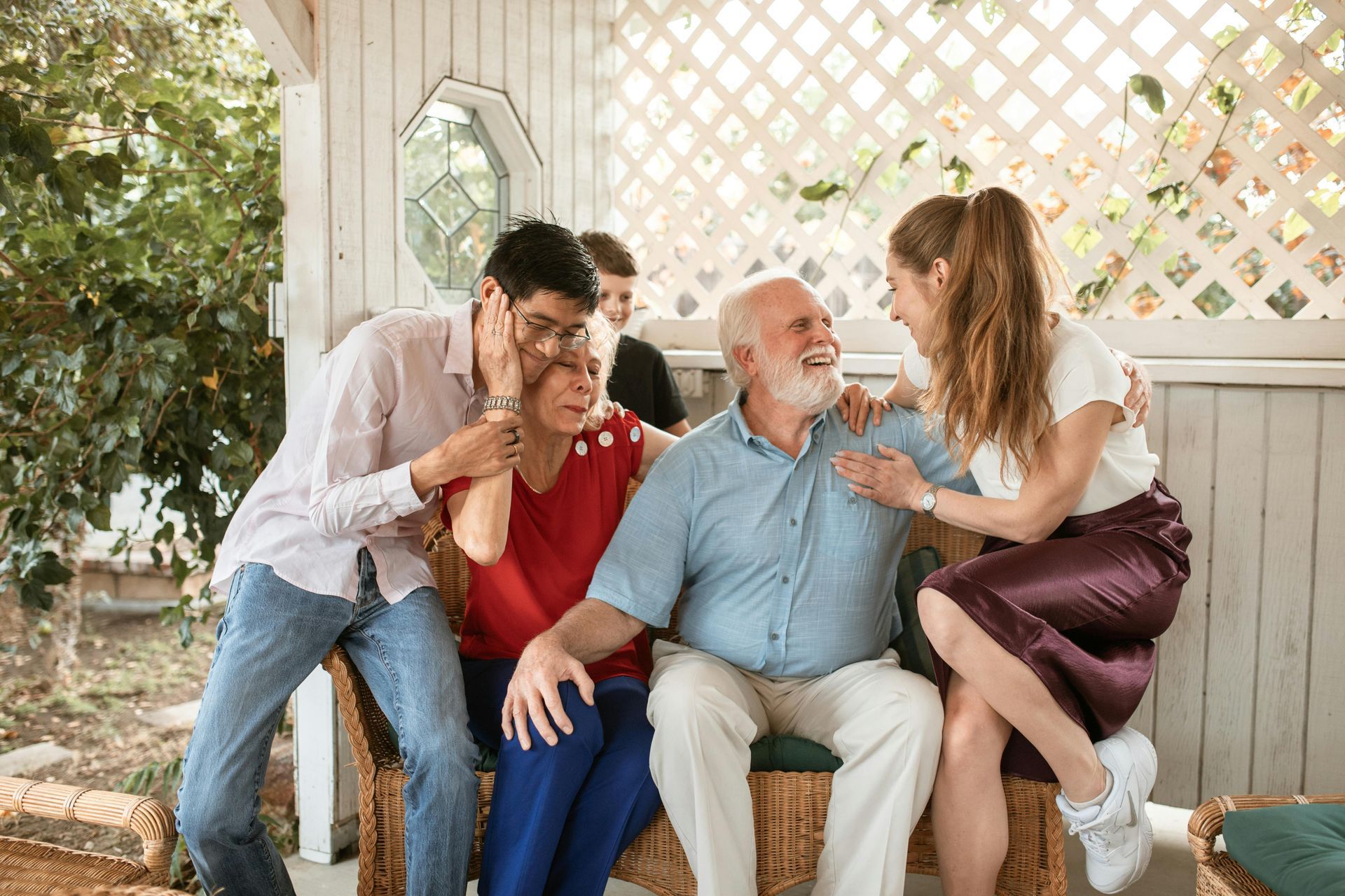 Family laughing on a porch. Elderly couple seated, surrounded by two younger adults who are embracing them.