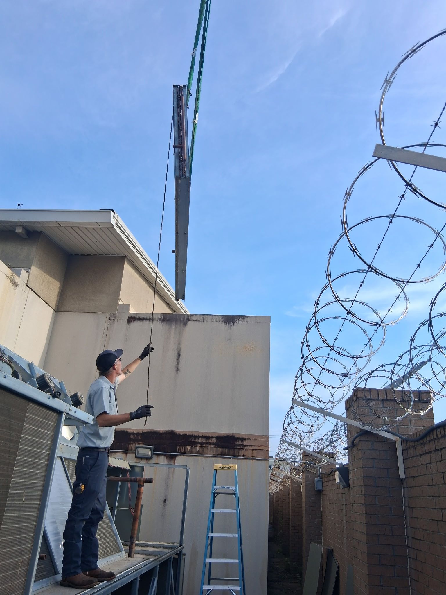 Worker on a ladder measuring a wall near rooftop barbed wire under a blue sky