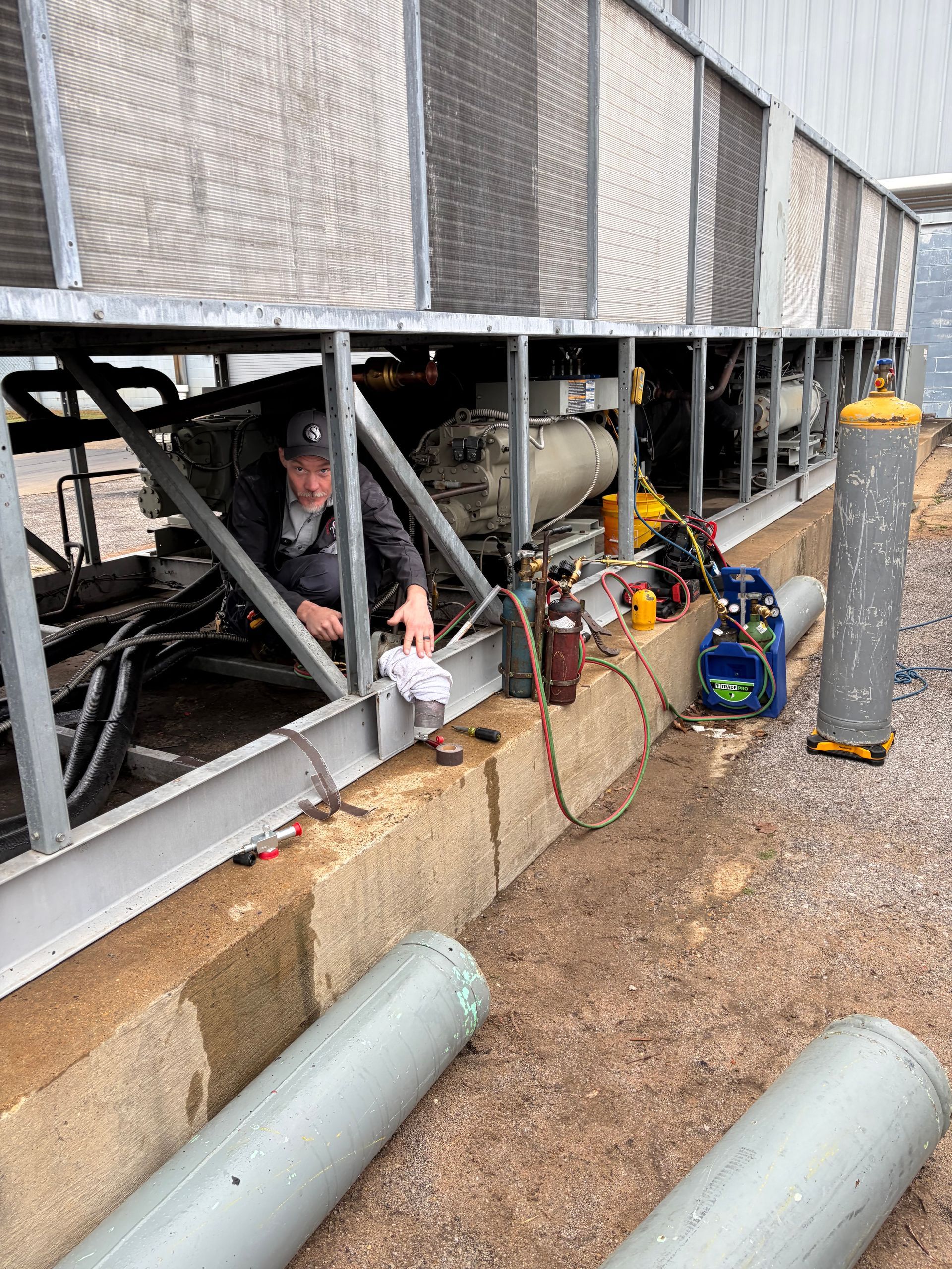 Technician working under rooftop HVAC units beside pipes and equipment on a wet gravel surface