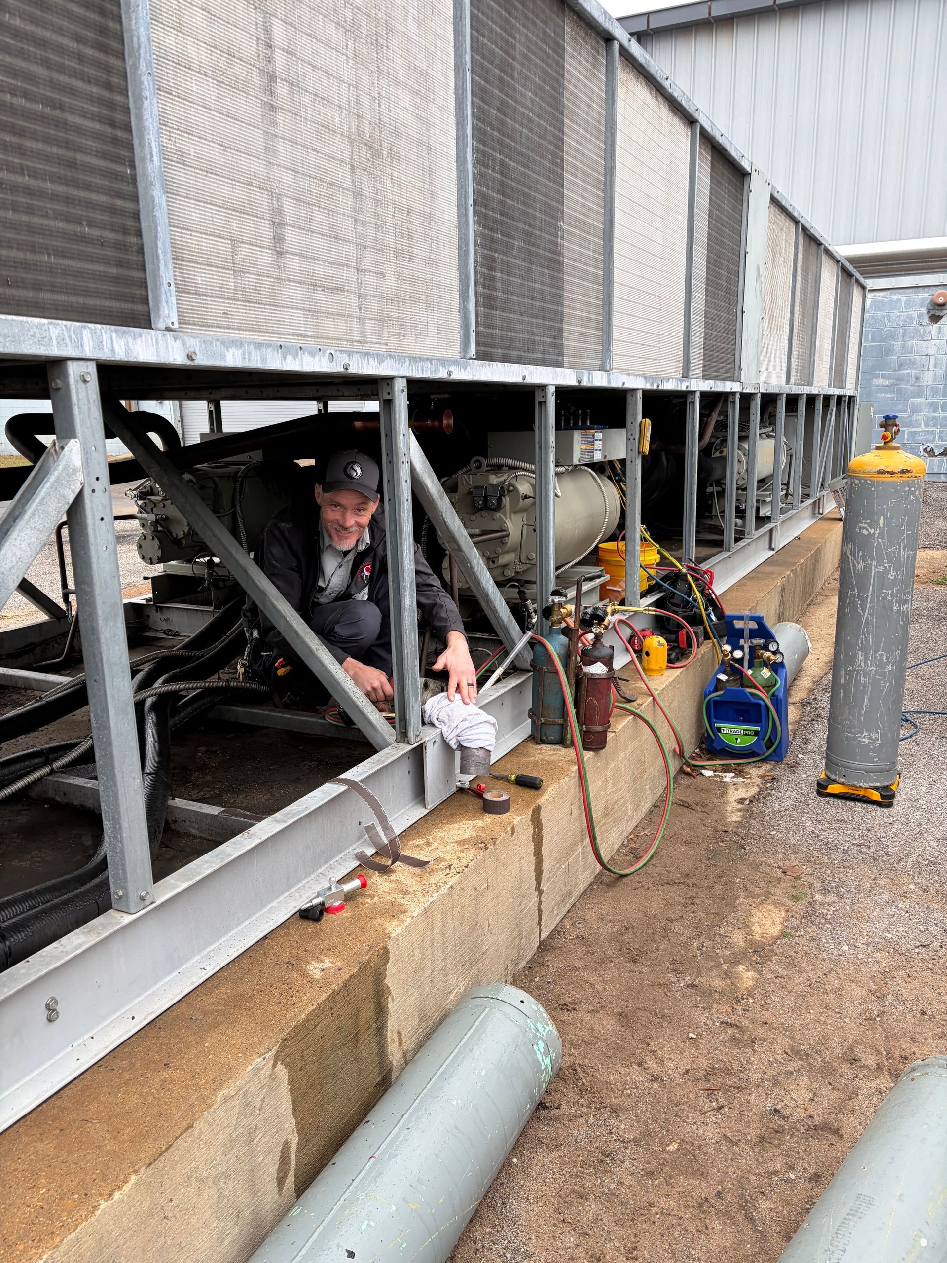 Worker crouches beneath metal trailer in a gravel yard, with tools and equipment nearby.