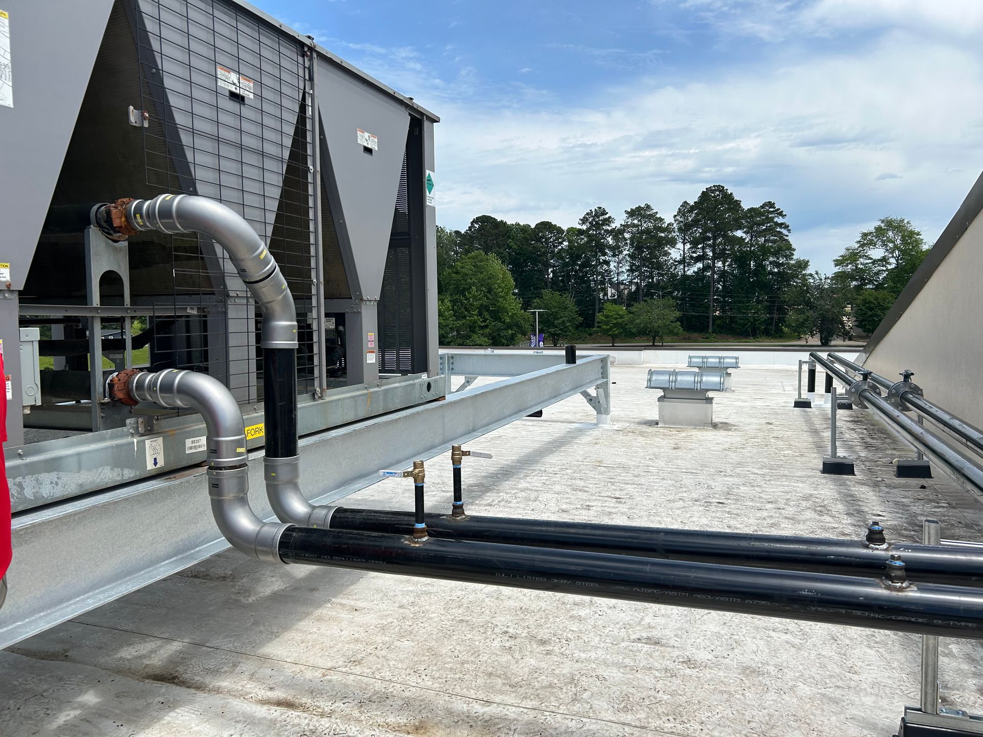 Rooftop HVAC piping and ducts beside a large industrial unit on a gravel rooftop.