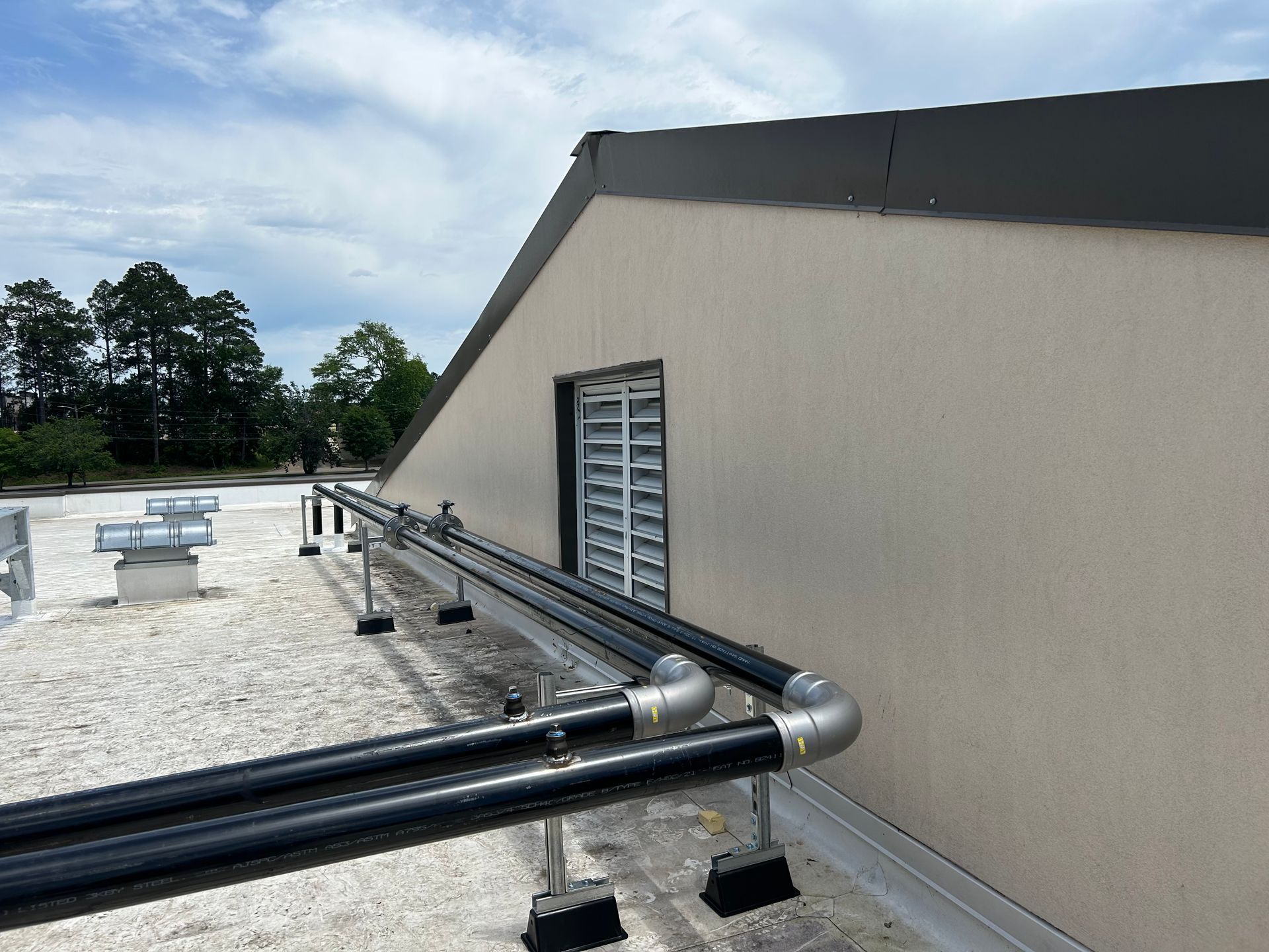Rooftop with black pipes along a beige wall, vented window, and cloudy sky over a gravel surface.