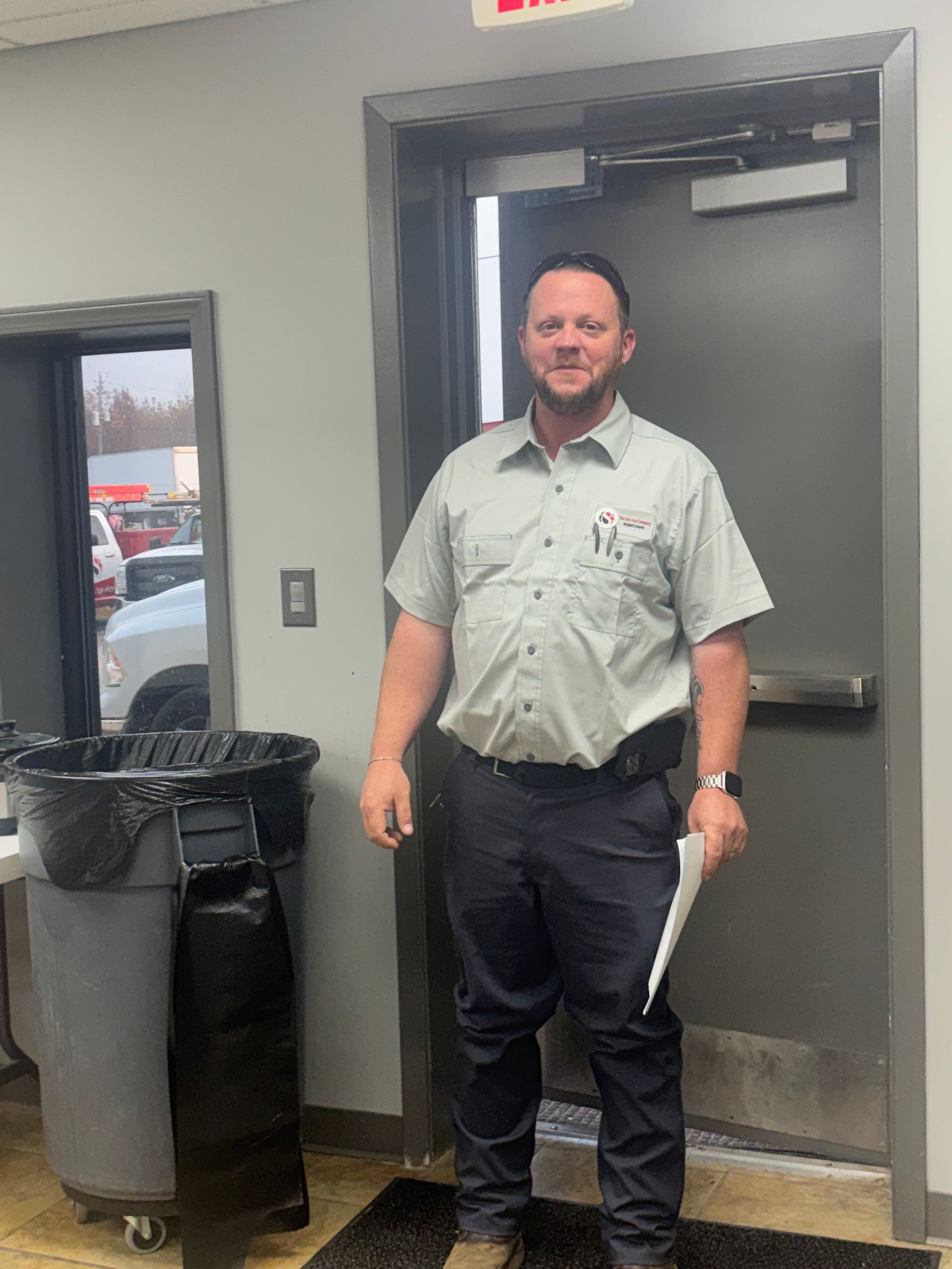 Man standing in a doorway holding papers, wearing a light shirt and dark pants beside a trash can.