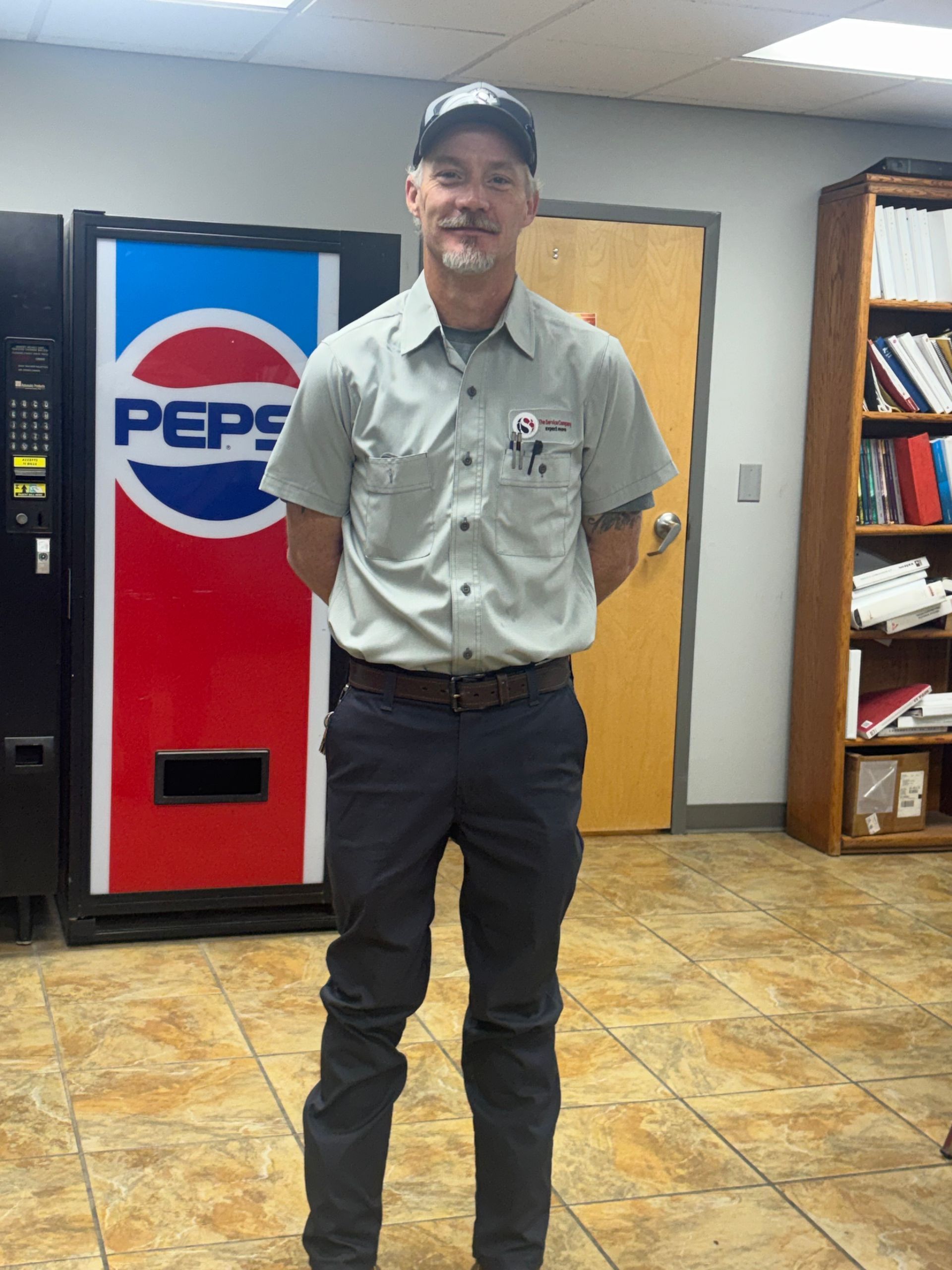 Man standing in an office break room near a Pepsi vending machine, wearing a cap and work clothes.
