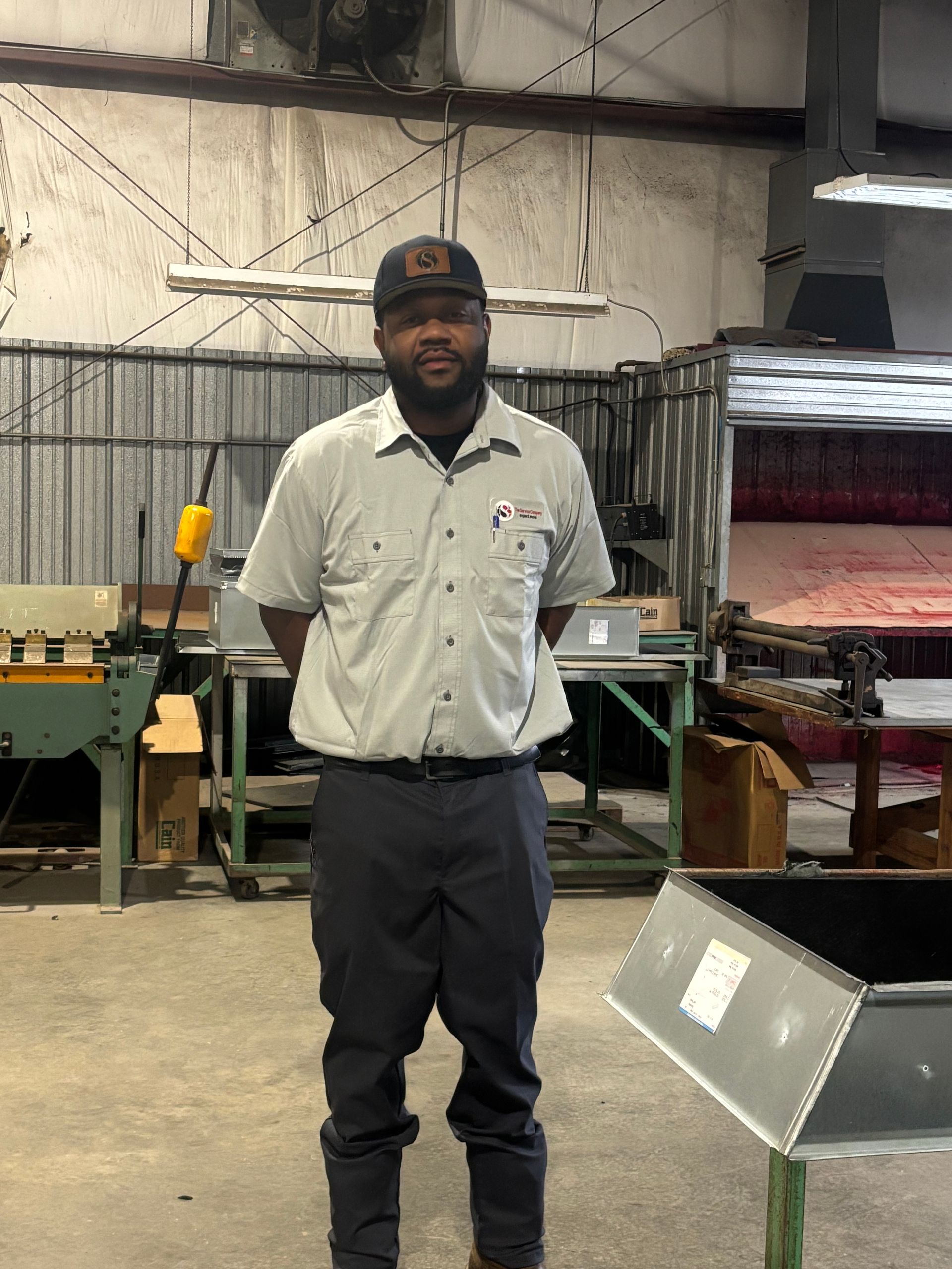 Man standing in a warehouse, wearing a light shirt and dark pants, with industrial equipment in the background.