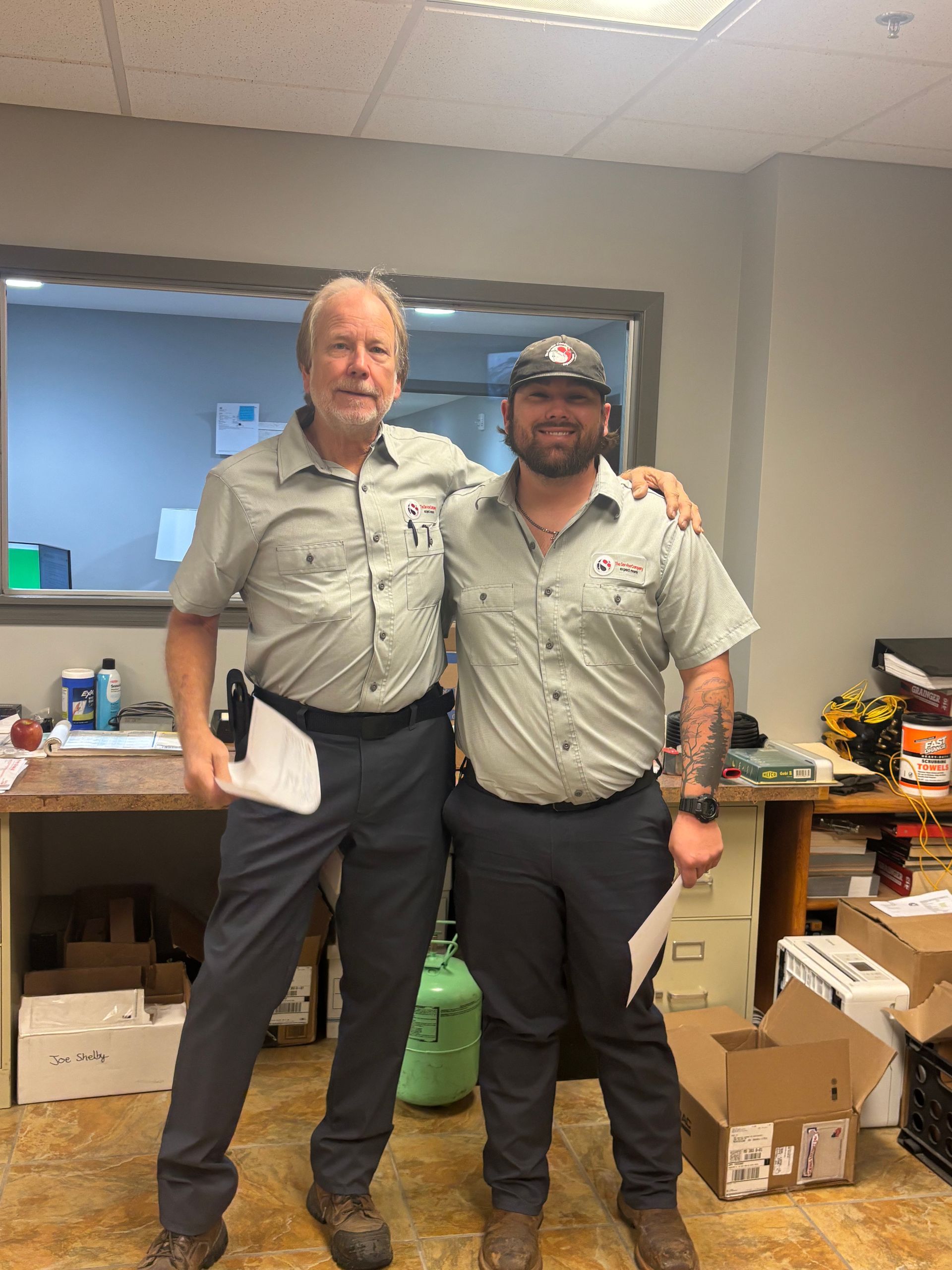 Two men in matching work shirts stand together in a break room, smiling with arms around each other.