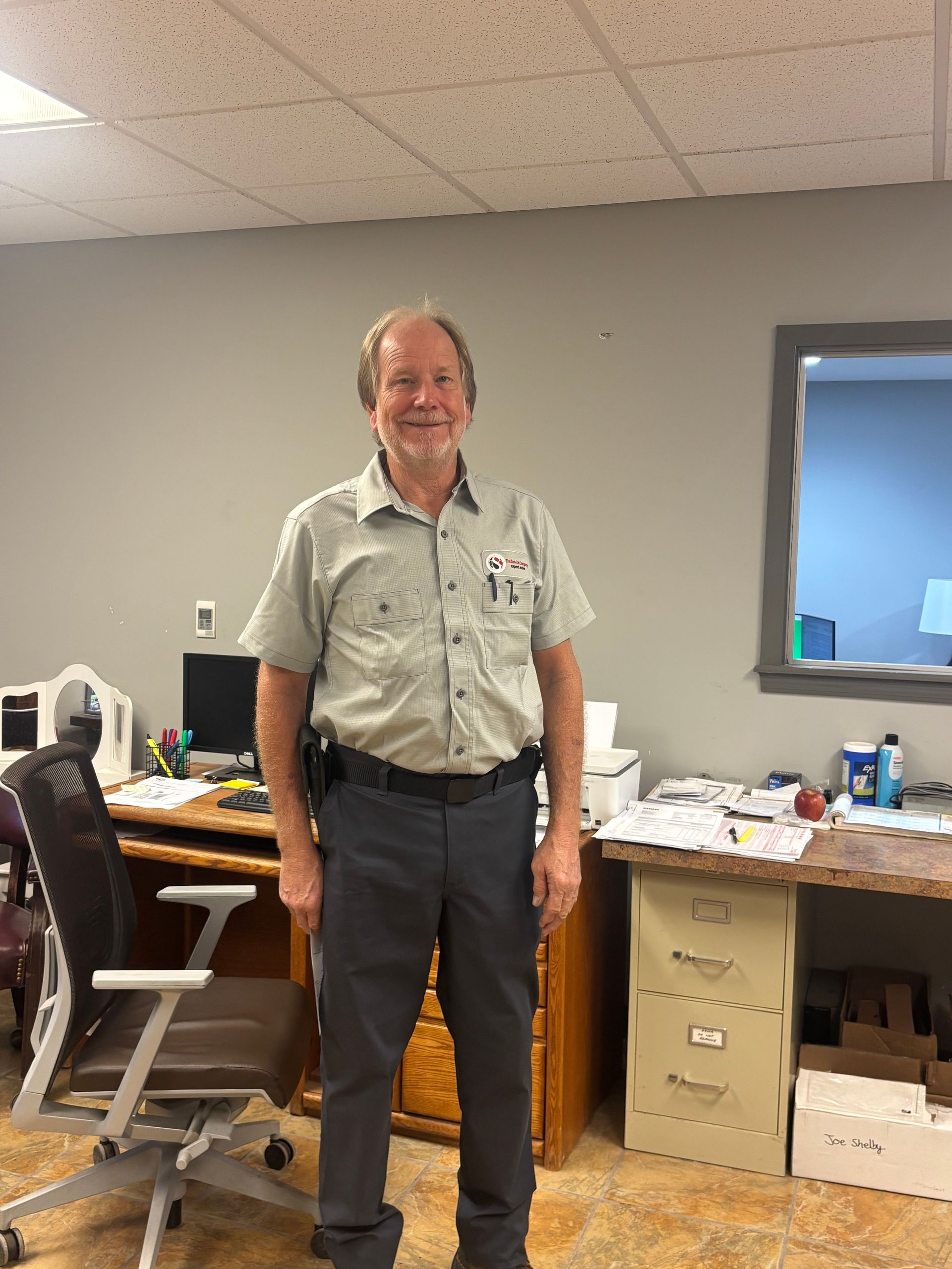 Man standing in an office beside a desk and chair, smiling at the camera.