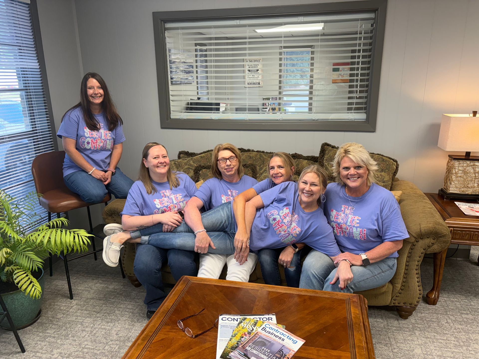 Six people in matching purple shirts pose in an office lounge, smiling around a couch and coffee table.
