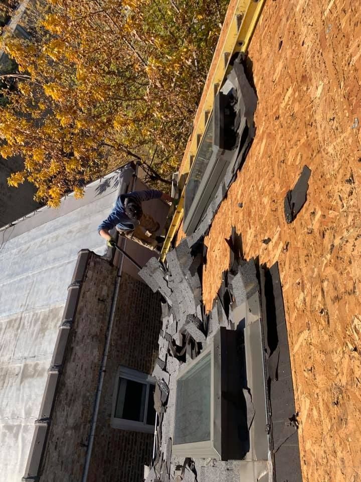 Collapsed rooftop debris beside a brick building and autumn trees, with shingles scattered below.