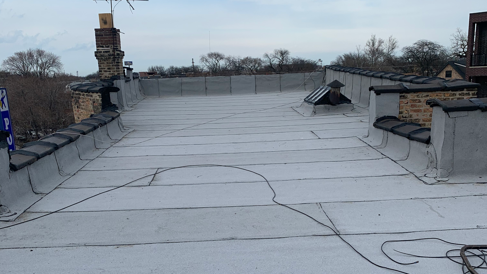Flat rooftop with white waterproof coating, vents, and a brick chimney under an overcast sky