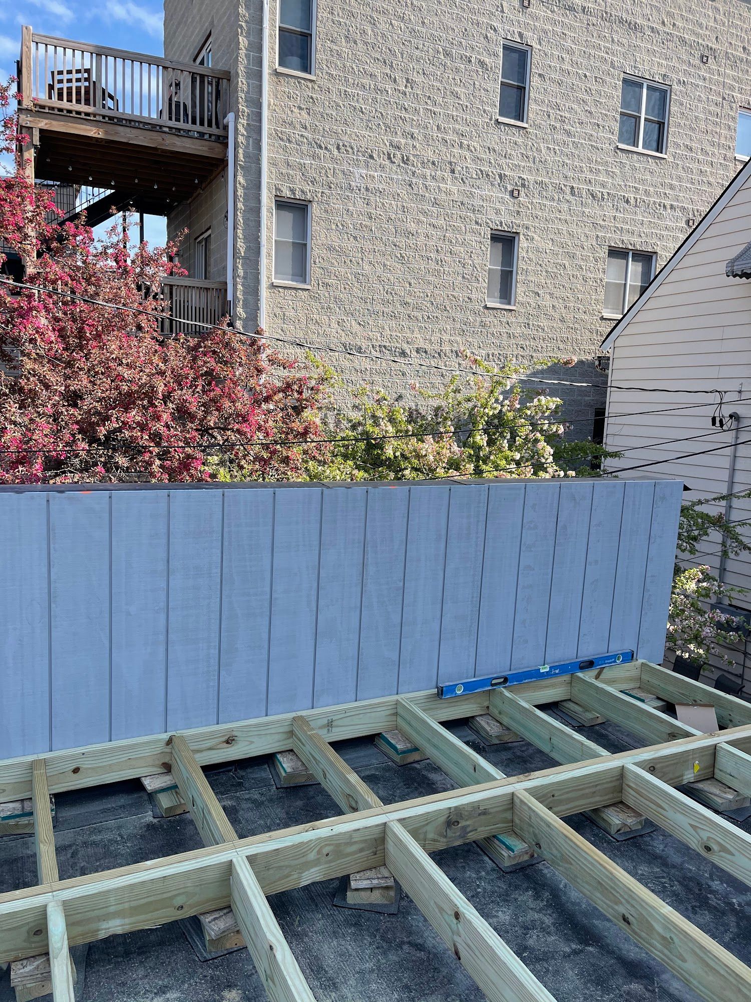 Backyard deck framing beside a blue fence, with trees and apartment buildings in the background