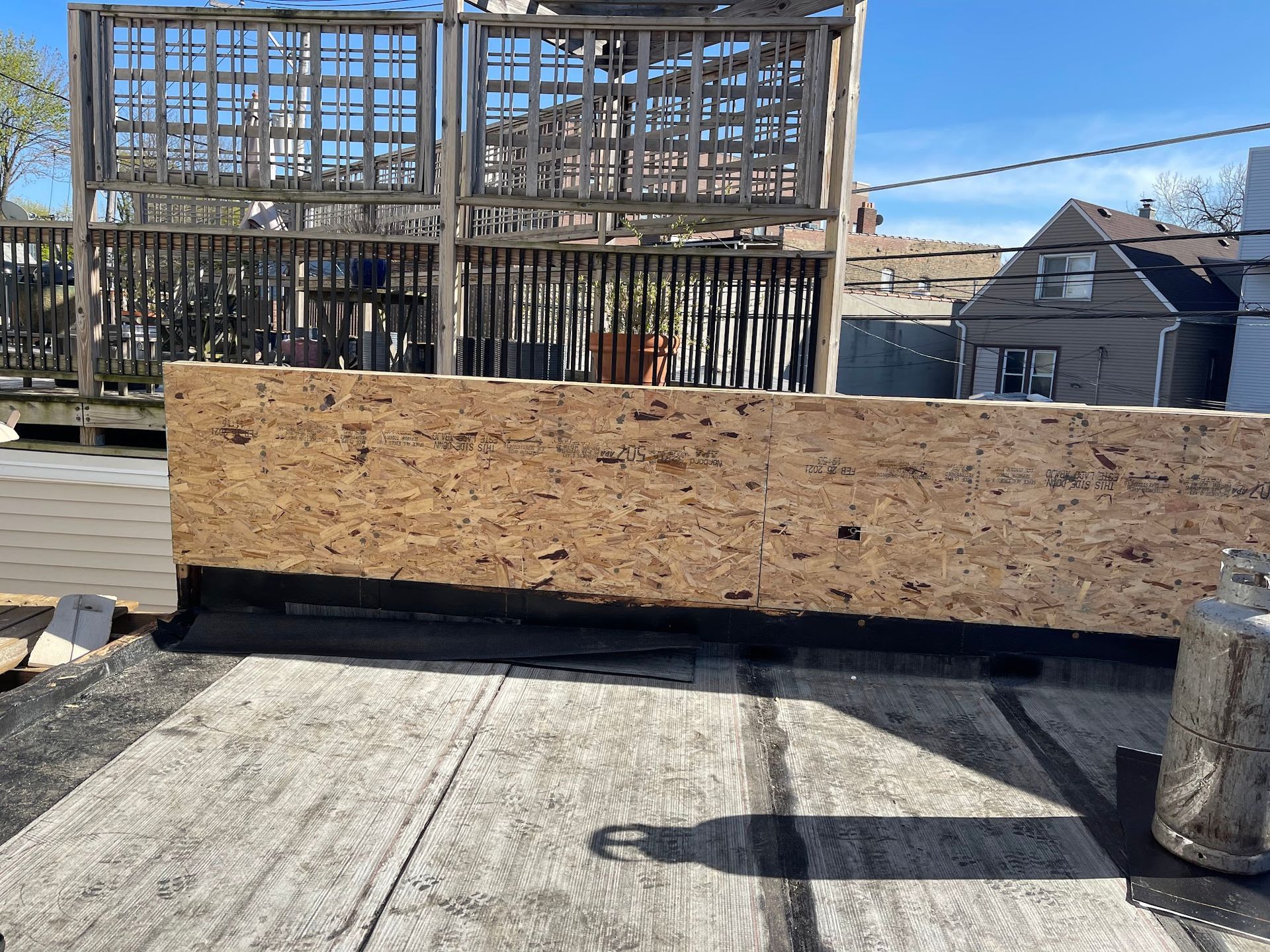 Rooftop construction site with plywood barrier, exposed framing, and neighboring houses in the background
