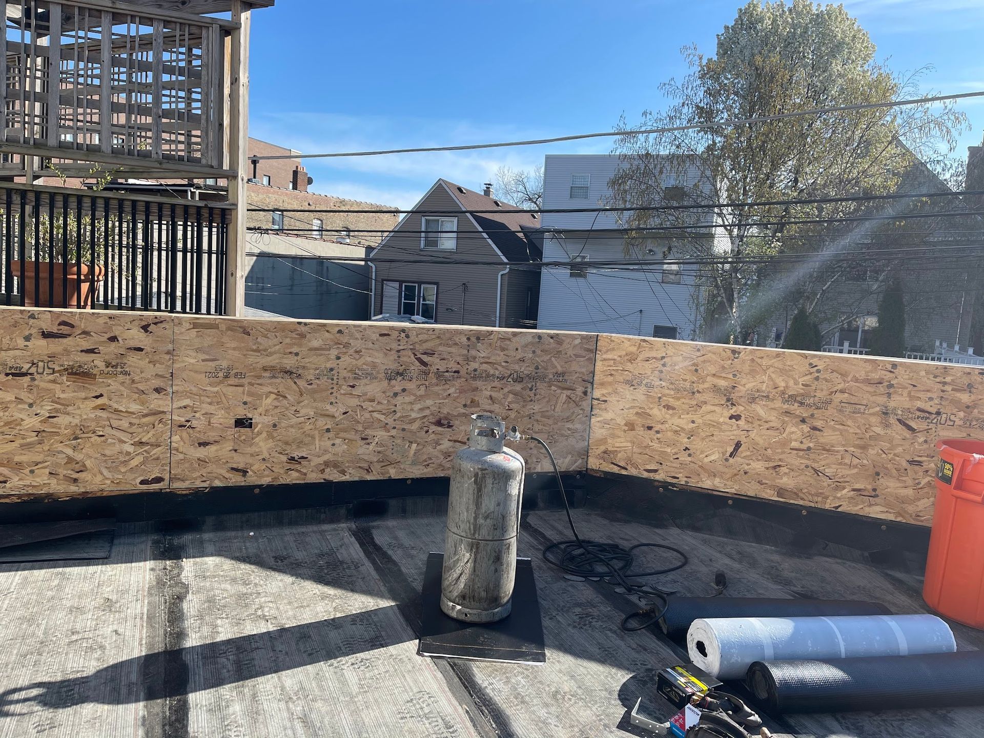 Rooftop construction area with plywood barriers, pipes, and buildings in the background under a blue sky