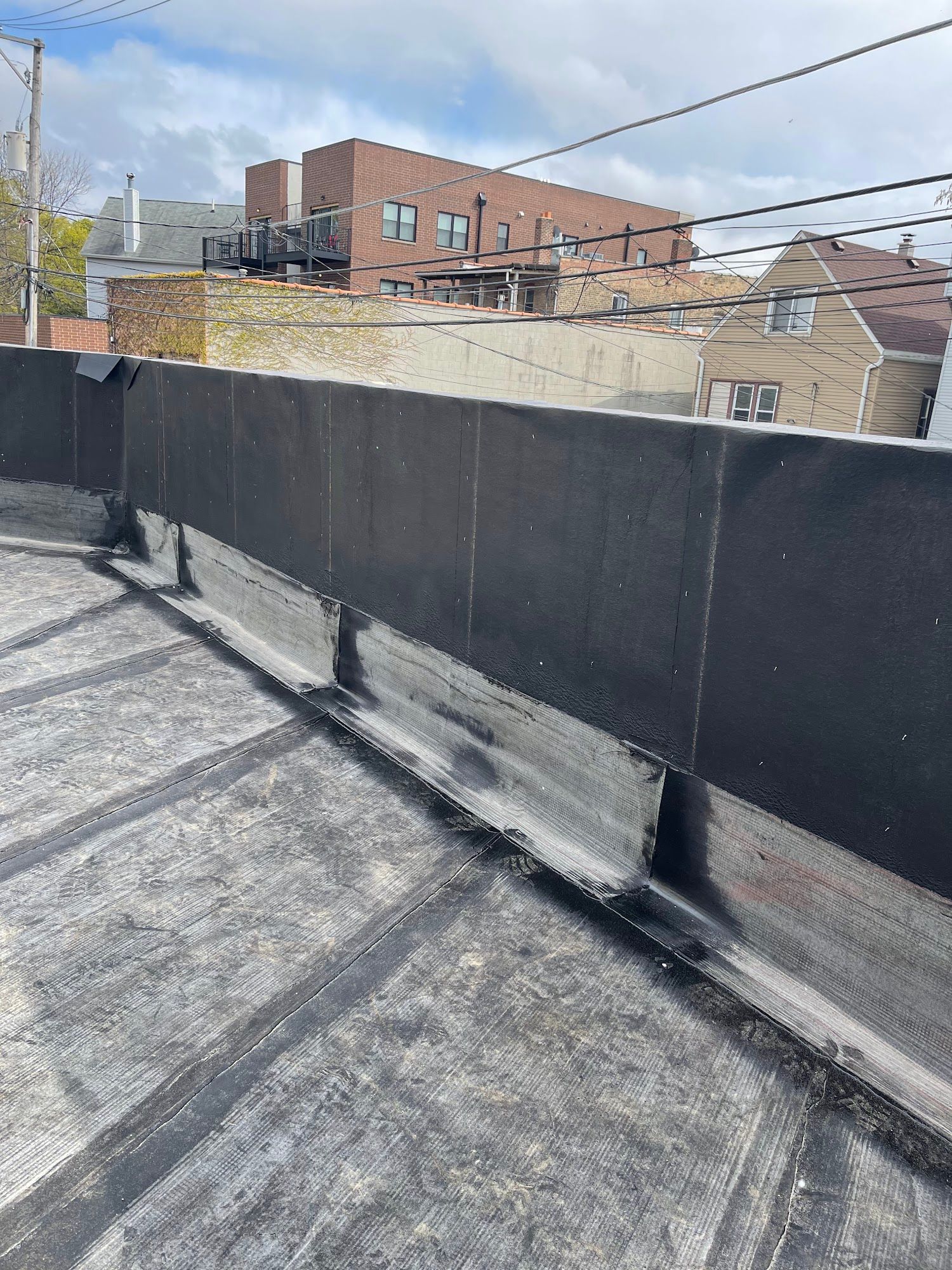 Rooftop with black parapet wall and rooftop equipment, overlooking brick buildings and power lines.