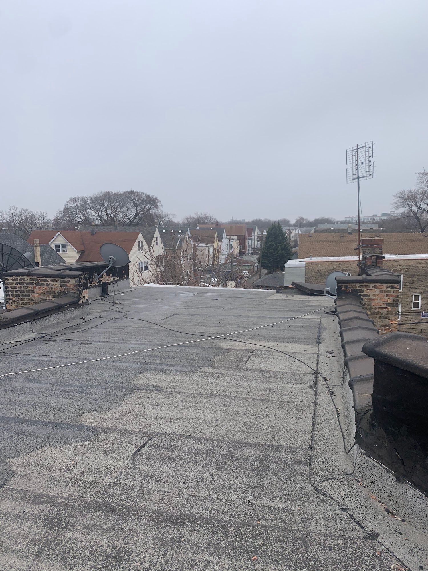 Rooftop with flat gray surface, stone parapets, and distant houses under an overcast sky.