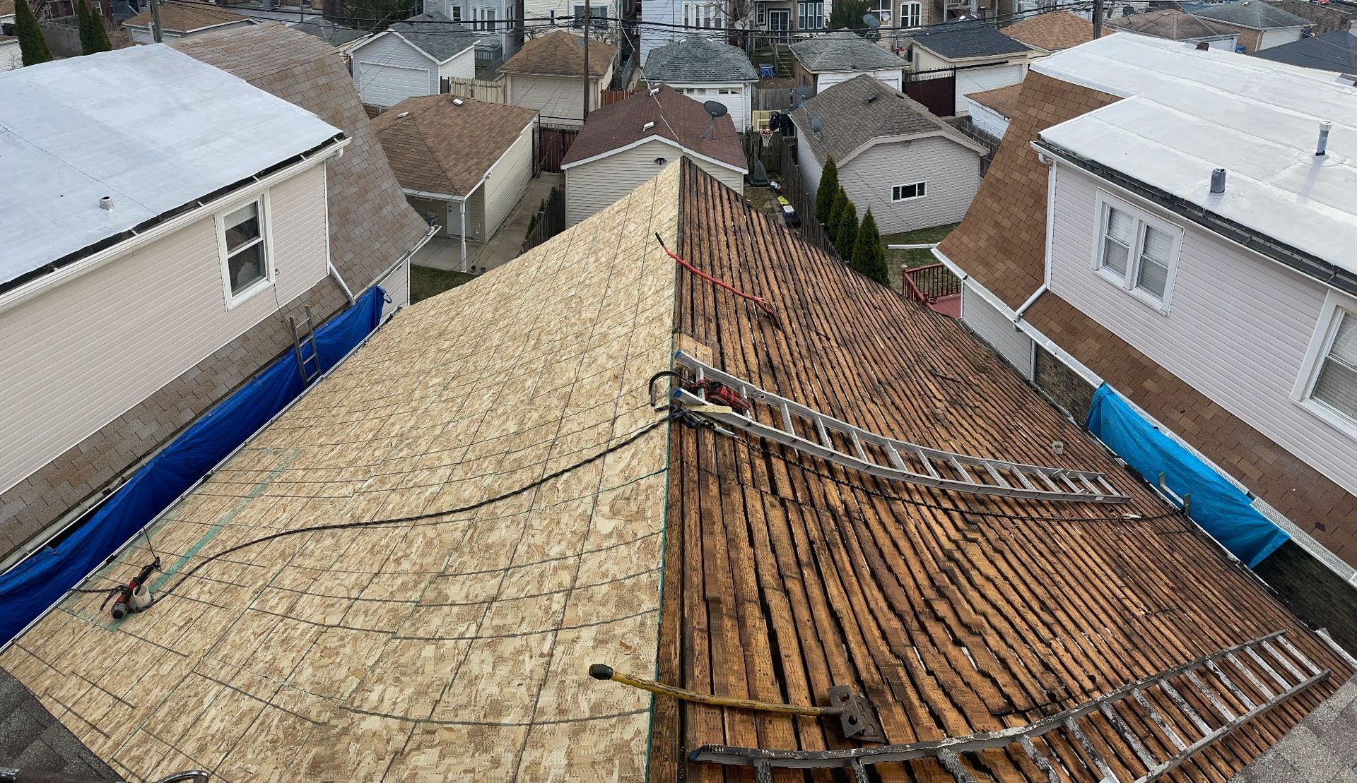 Rooftop under repair with new plywood on left and old clay tiles on right, viewed over a neighborhood.