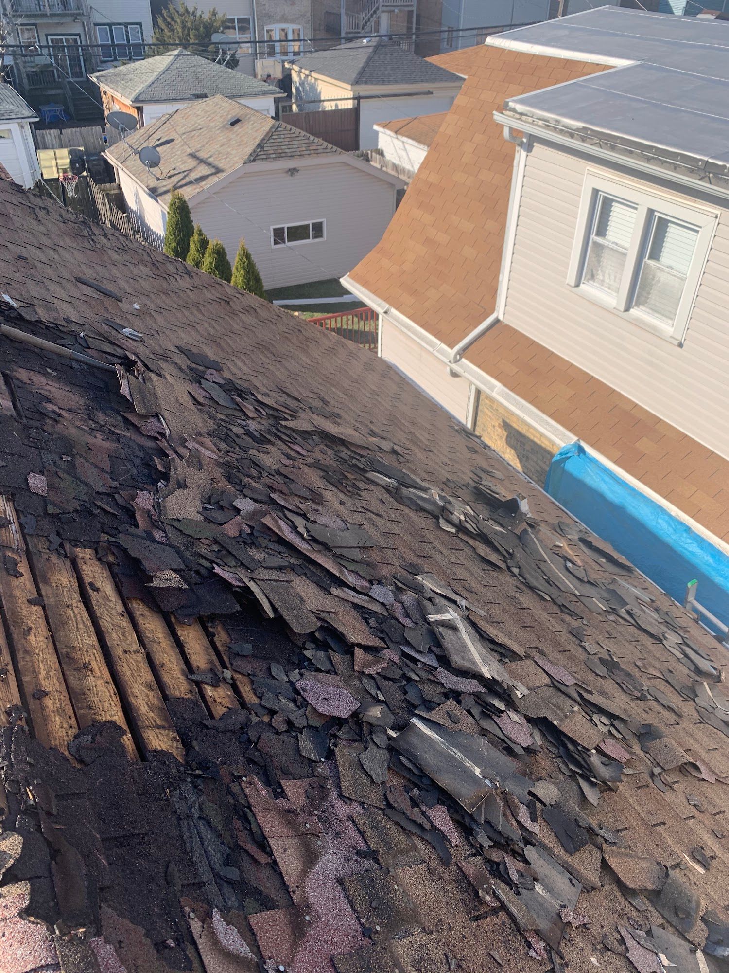 Damaged shingled roof in foreground overlooking neighboring suburban houses on a sunny day