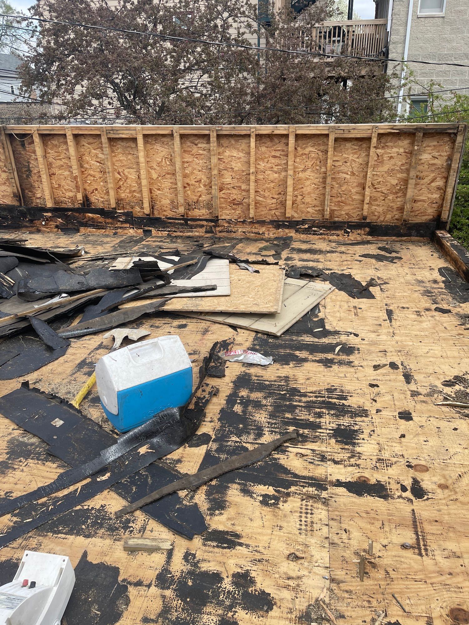 Damaged rooftop with torn black roofing, exposed plywood, and a blue-white cooler near the wall.