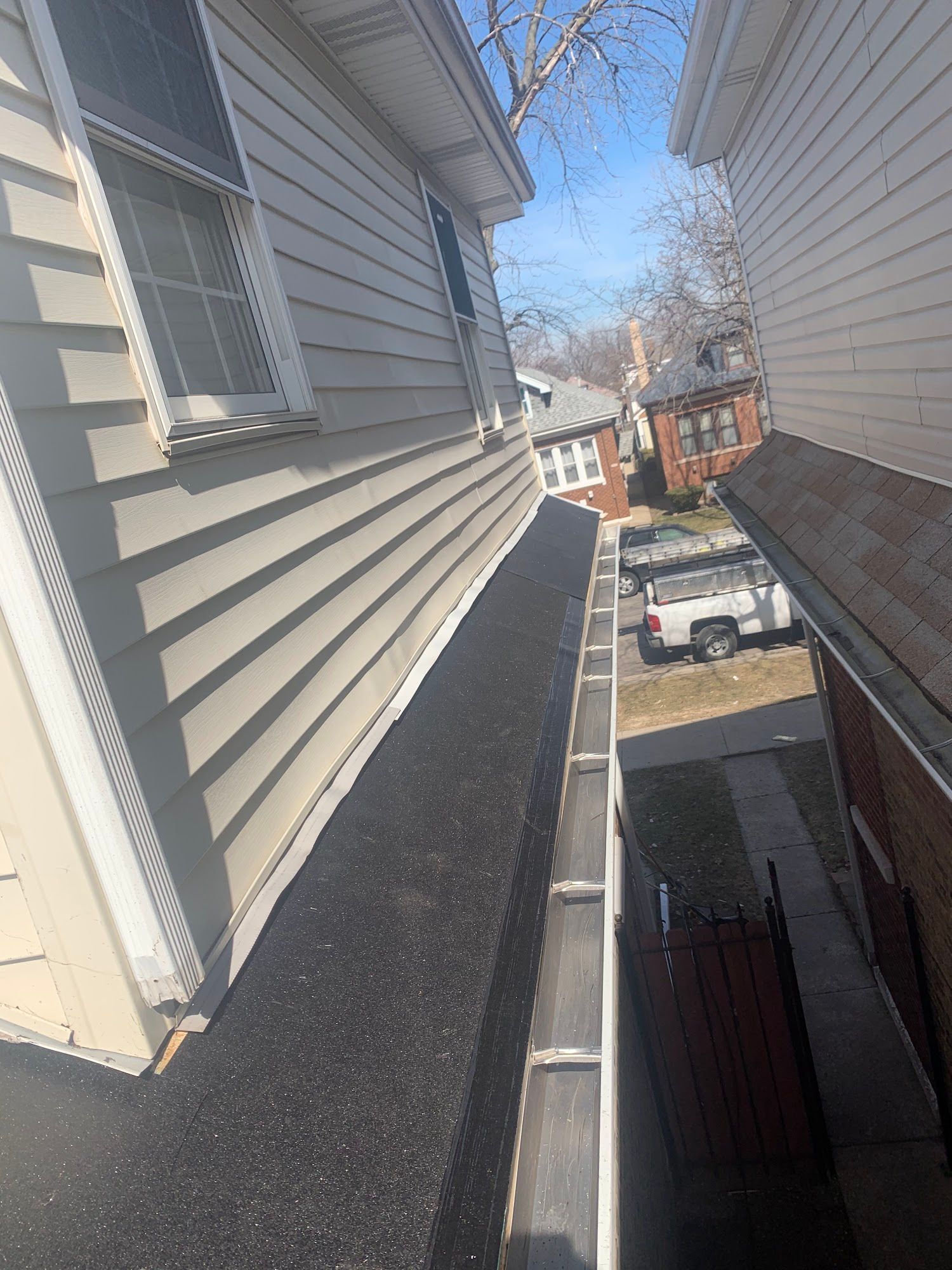 Narrow rooftop walkway beside gray siding, overlooking a residential street and parked cars.