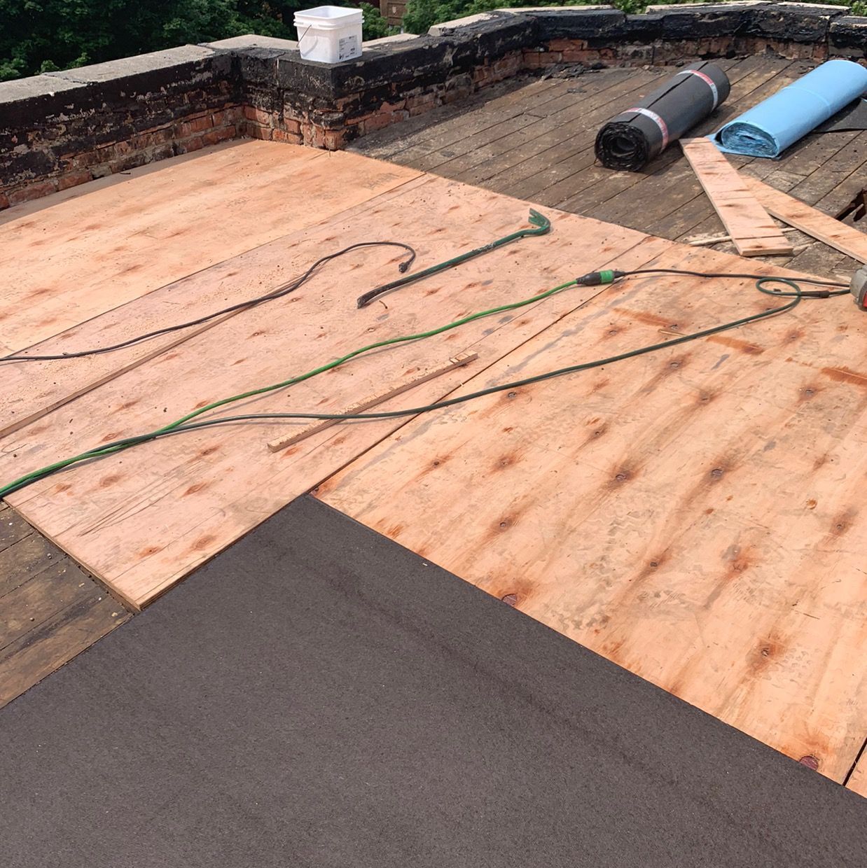 Roofing work in progress on a flat roof with plywood, black underlayment, and rolls of roofing material