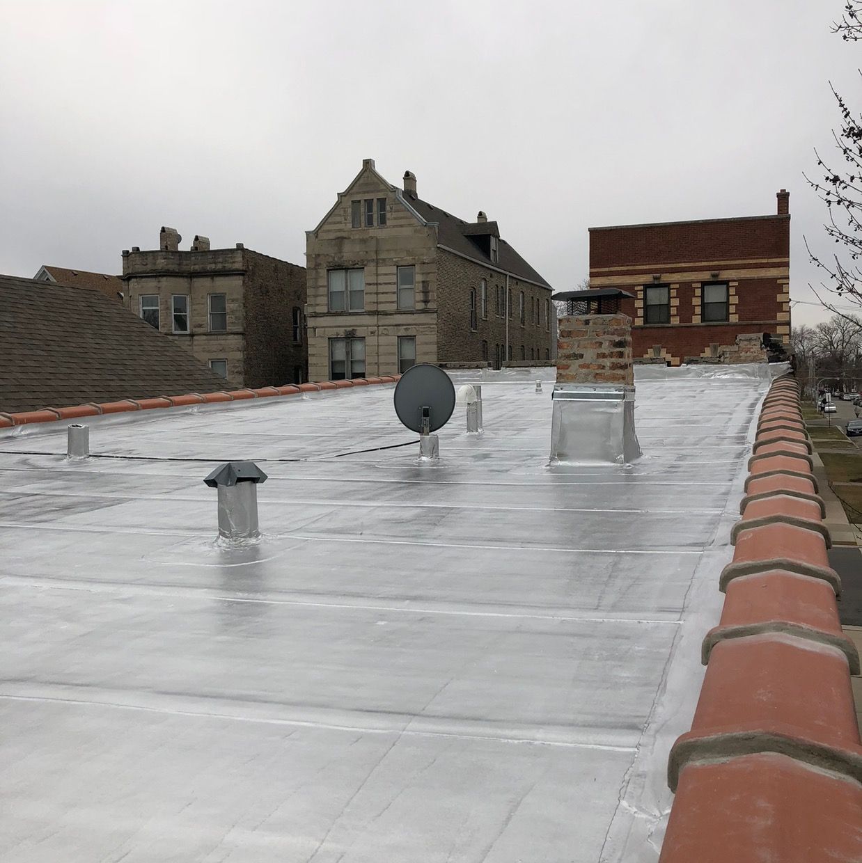 Rooftop with silver waterproofing, vents, and a satellite dish beside brick buildings under an overcast sky