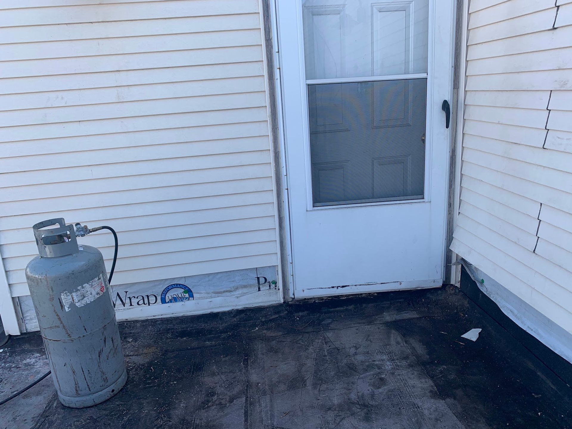 Propane tank beside a white exterior door and siding on a dark porch.