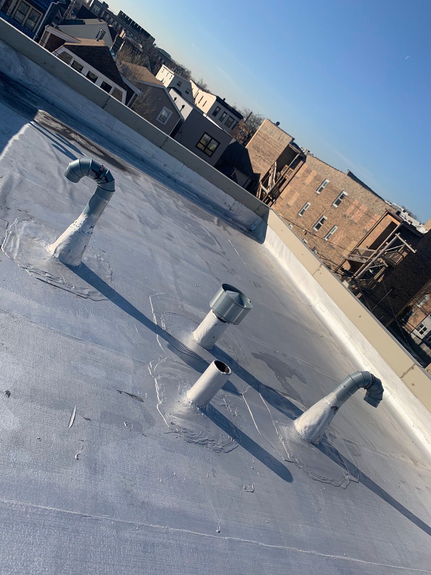 Rooftop with white coating, vent pipes, and a city skyline under a clear blue sky.