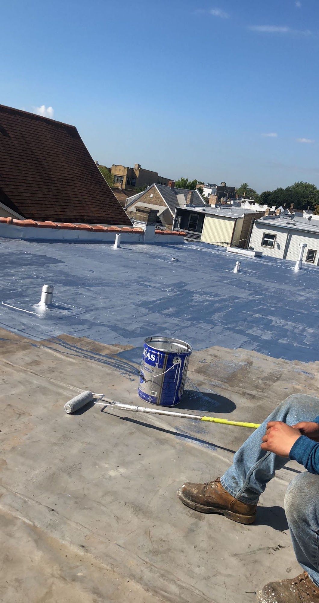 Rooftop with blue waterproofing membrane, paint can and roller, and a worker’s boots at right edge