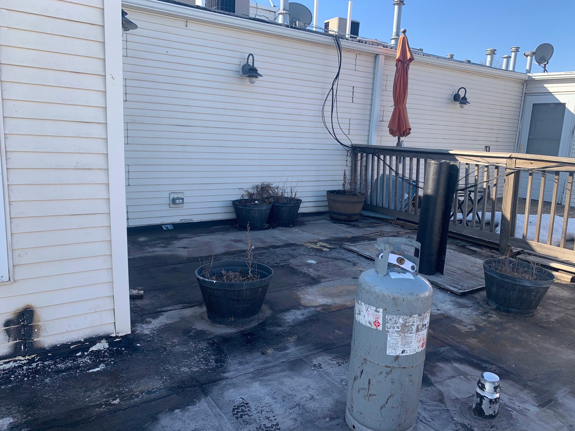 Rooftop patio with potted plants, a closed red umbrella, and a small grill beside a white wall.